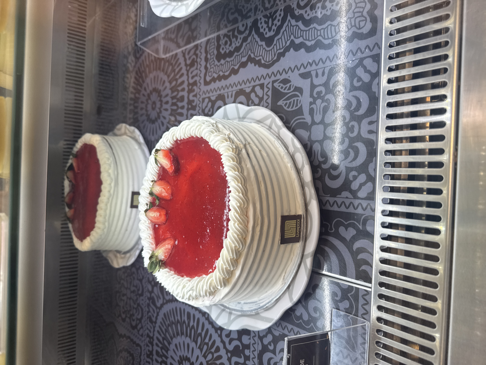 The image shows two strawberry cakes displayed in a refrigerated pastry case. The cakes are round with white frosting and a red strawberry topping, adorned with fresh strawberries. The cake on the right has a "Liverpool" logo. The display case is located in a Liverpool department store in Puebla, Mexico. The cakes sit on white plates, and the display case has a patterned backdrop.