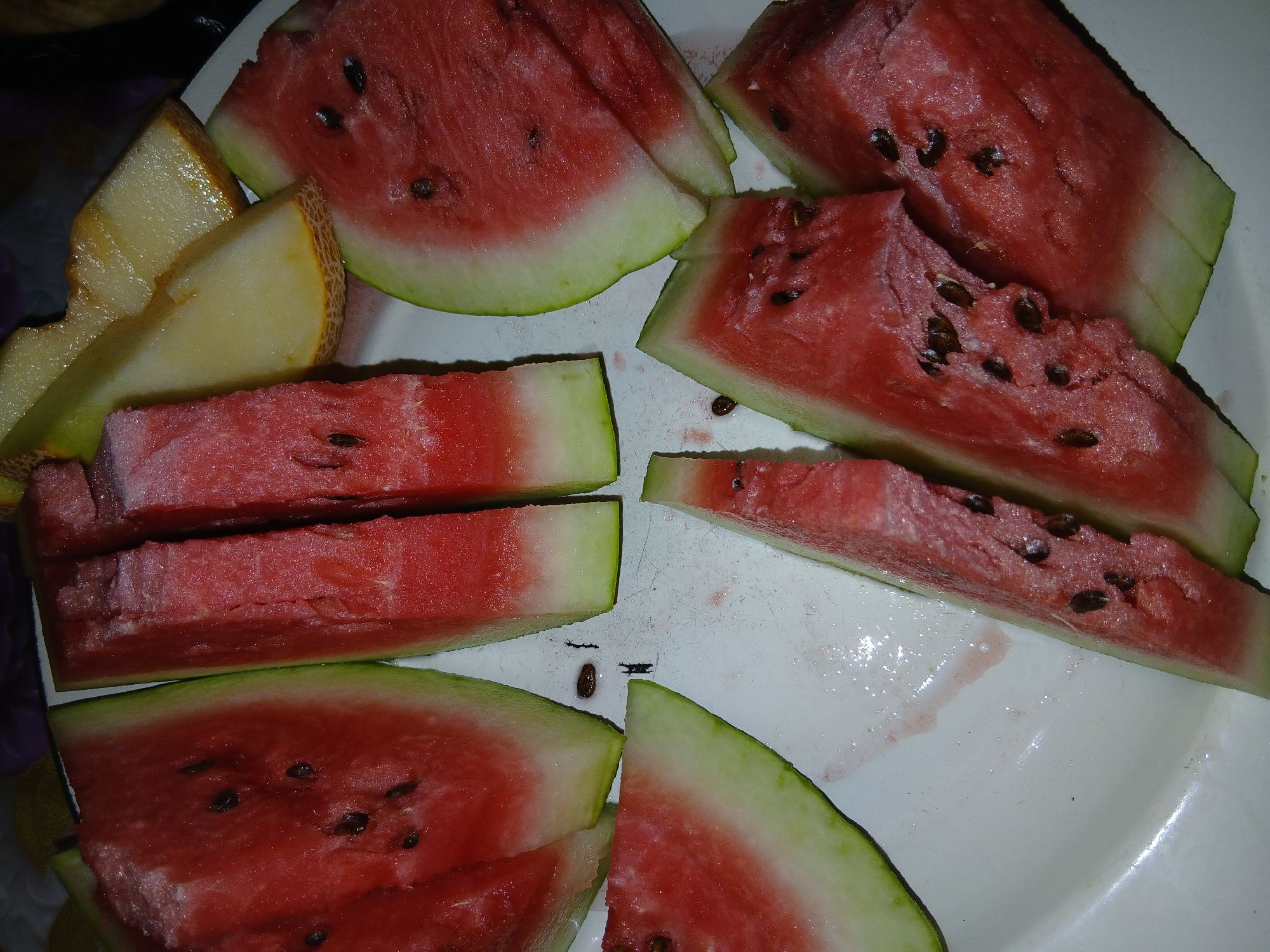 A top-down close-up shot shows a plate of sliced watermelon and cantaloupe. The watermelon is cut into wedges with the rind still attached, and many of the seeds are visible. Some pieces of cantaloupe, with their rind on, are interspersed among the watermelon. The fruits are arranged on a white plate, which has some juice stains and a few dark specks. The background is dark and out of focus, suggesting indoor lighting. The overall impression is a casual, refreshing dessert.
