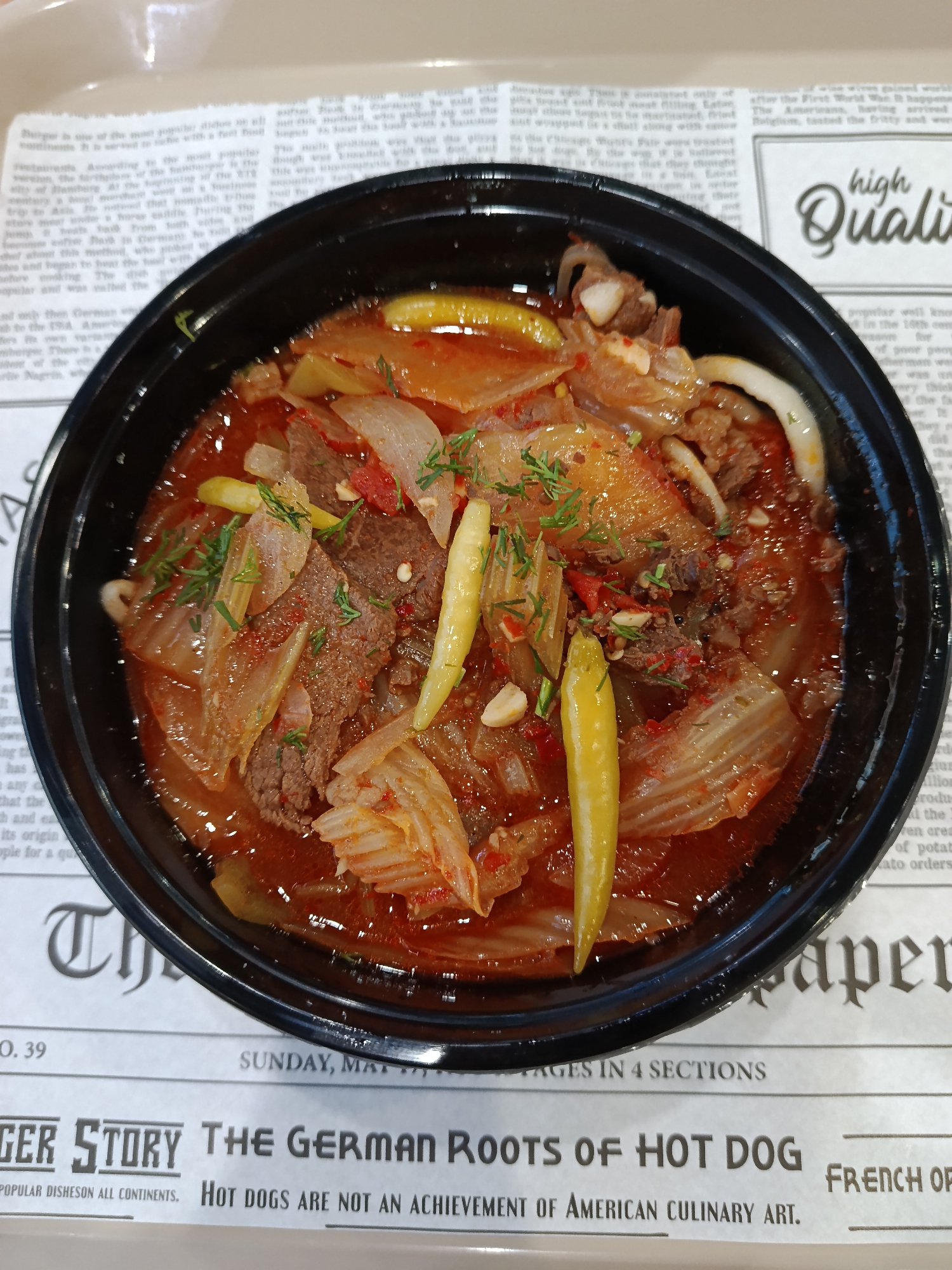 A close-up shot shows a black bowl filled with a red, spicy stew, possibly kimchi jjigae, sitting on a newspaper-printed surface. The stew contains beef, onions, kimchi, and yellow peppers, garnished with dill. The newspaper features headlines about the origins of hot dogs. The location, Moscow, Russia, is not visually apparent in the image.