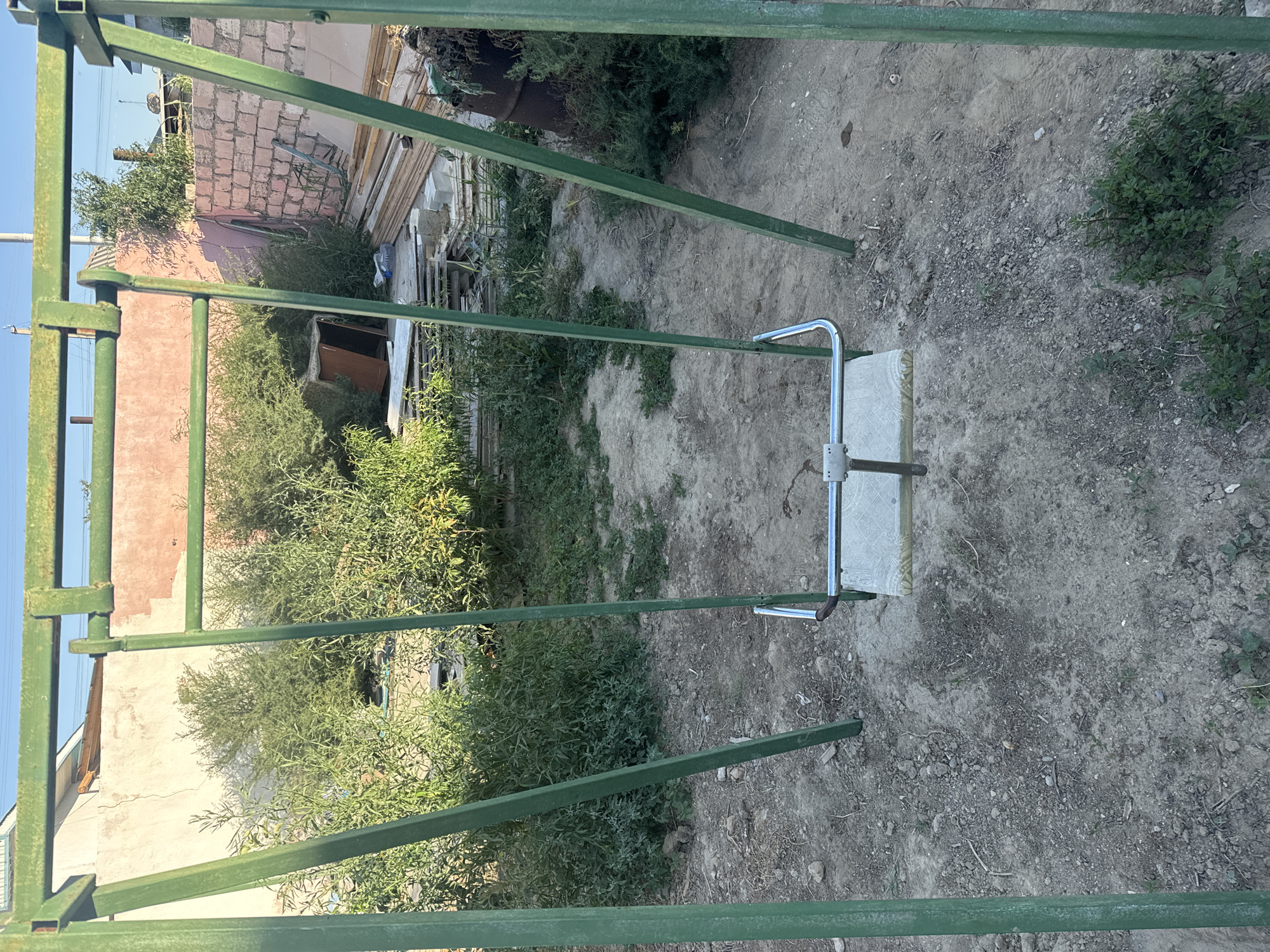 The image shows a green metal swing set in what appears to be a yard or outdoor area. The swing set has a single seat, which is a rectangular platform with a metal bar across the front. The ground around the swing set is dirt and gravel, with some scattered rocks and sparse vegetation. In the background, there is a pinkish-colored wall, some overgrown bushes and trees, and stacks of wood and building materials. There are also some discarded items visible, including a dark barrel and what looks like a piece of furniture. The overall impression is one of a somewhat unkempt outdoor space, possibly in a residential area. The location is identified as Atameken, Kazakhstan. There are no people or animals visible in the image.
