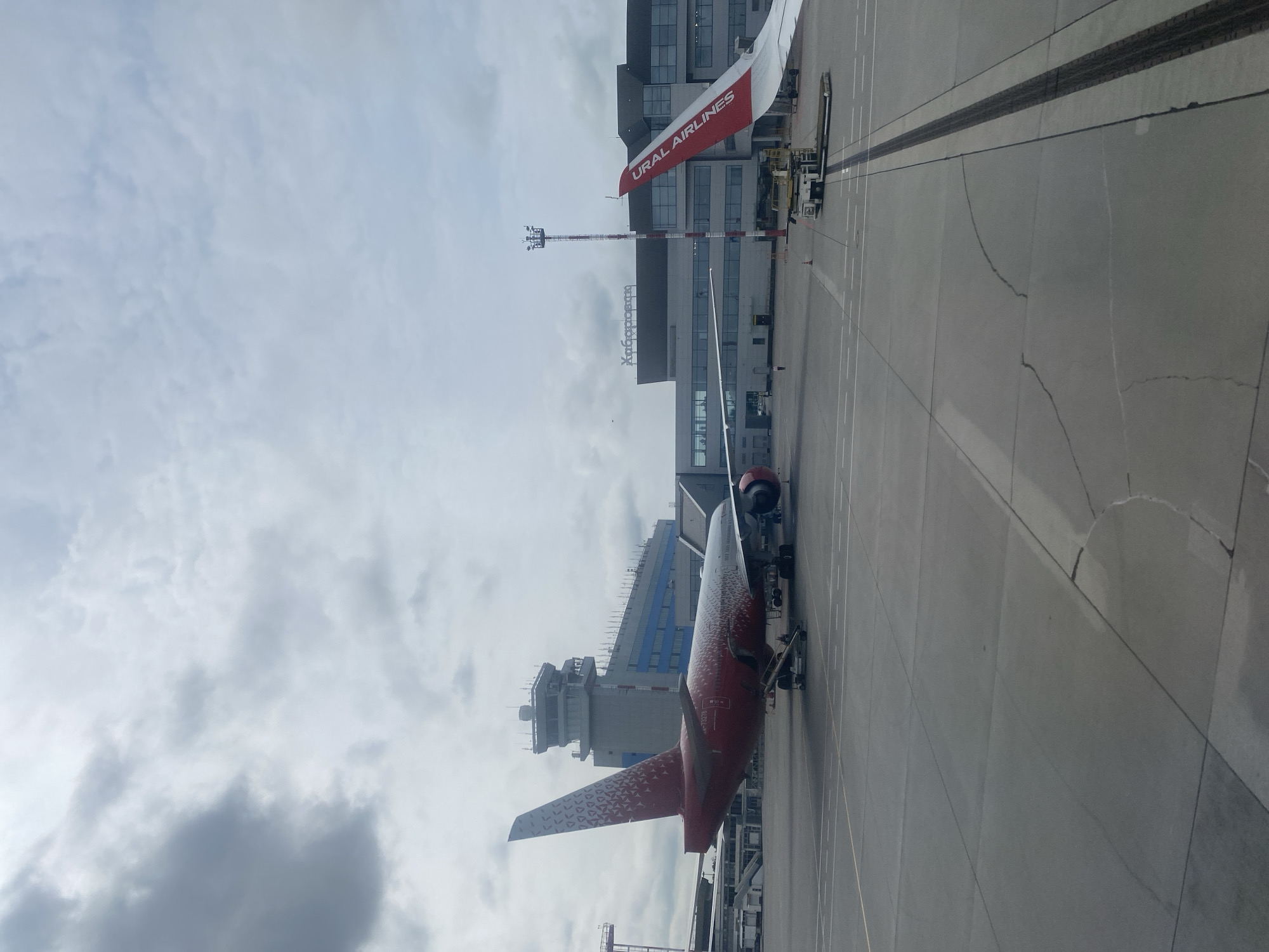 An exterior, medium-shot captures an airport tarmac at an unspecified time of day. In the foreground, the wing of a red and white airplane, emblazoned with "URAL AIRLINES," juts into the frame from the right. The plane's wing is white with a red stripe at the top, and the airline's name is written in red lettering. To the left, a red airplane with a white tail is parked on the tarmac, facing left. The tail of this aircraft is adorned with a white, repeating pattern of small triangles and circles. The fuselage is predominantly red with similar white patterns. The plane is connected to a mobile staircase. In the background, a modern airport terminal building with large windows is visible. Above the terminal, signage reads "Хабаровск" (Khabarovsk). A control tower with multiple levels and antenna structures stands tall to the left of the terminal. Several vehicles, including a baggage tug, are positioned on the tarmac near the terminal. The sky is overcast with a mix of dark and light clouds. The ground is paved with concrete, marked with painted lines and exhibiting some cracks. The scene appears to be at the Khabarovsk Novy Airport, serving the city of Khabarovsk, Russia. No individuals are clearly discernible.