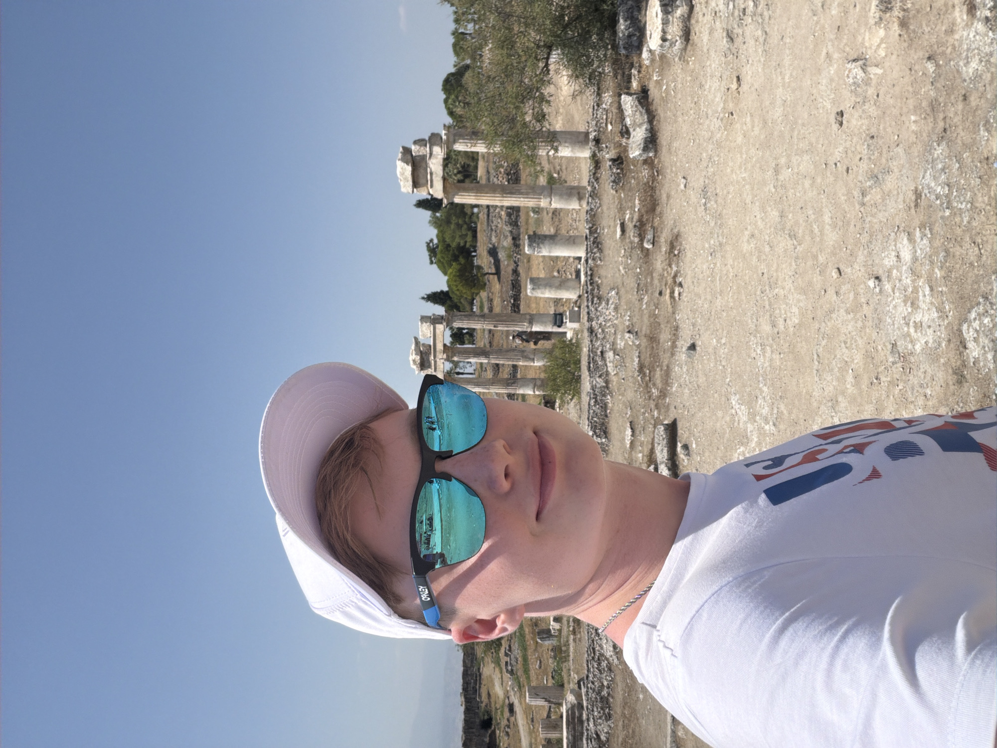 This is a selfie of a young Caucasian male wearing a white baseball cap and bright blue mirrored Oakley sunglasses. He is wearing a white t-shirt with a red, white, and blue graphic on the left side of his chest and a silver chain around his neck. He is standing in front of ancient ruins under a clear blue sky. The ruins consist of weathered stone columns and debris, suggesting an archaeological site. In the background, trees and more scattered ruins are visible. The ground is a dry, dusty earth mixed with small stones and larger rock fragments. The person is smiling slightly at the camera. The scene appears to be an outdoor historical or archaeological site in Pamukkale, Turkey, possibly Hierapolis.