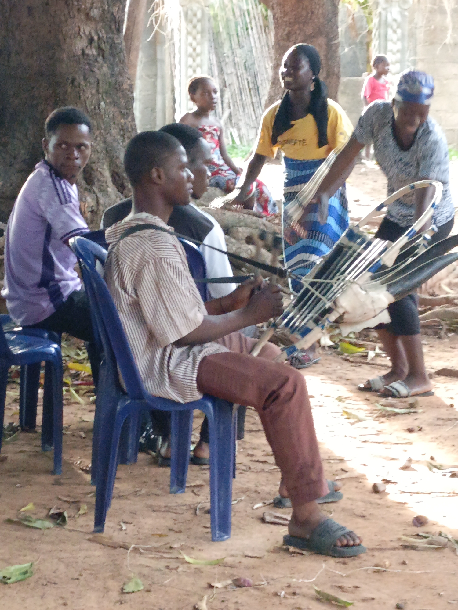 Akwanga, Nigeria: Several individuals are gathered outdoors in a sandy area with trees and a partial view of a fence in the background. A man in a light purple shirt is seated on a blue plastic chair and is looking toward the camera. Another man in a striped shirt and brown pants is seated on a similar blue chair; he is holding what appears to be a harp-like instrument. An older woman with a blue head covering and patterned shirt is holding the instrument. A woman in a yellow shirt and blue skirt stands nearby. In the background, another child can be seen. The scene appears to be a casual gathering.