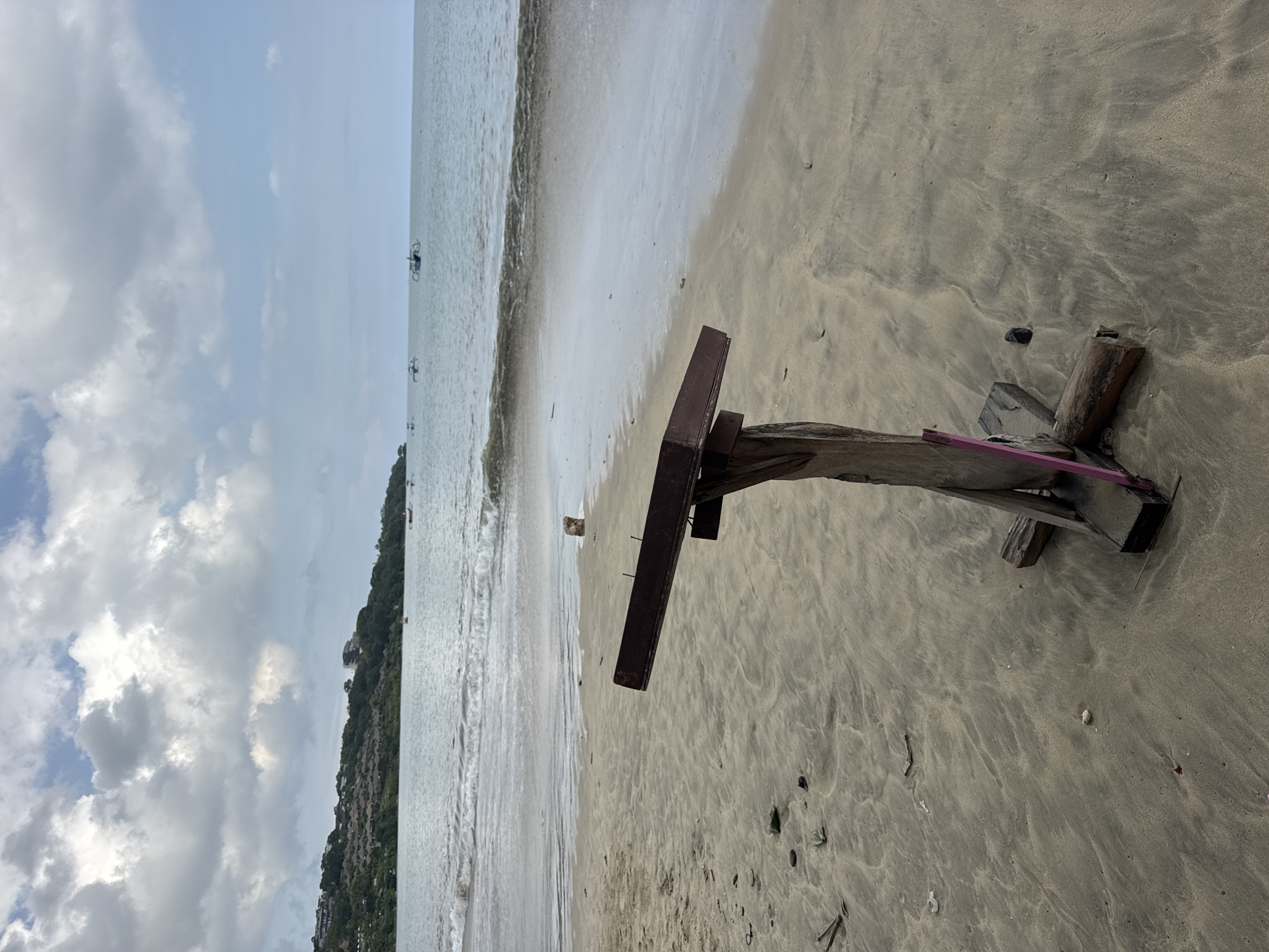 The image depicts a beach scene, likely in Kuta Selatan, Indonesia, given the geographical context. A weathered wooden structure, possibly the remnants of a beach chair or small table, lies abandoned on the sandy shore. The beach itself shows signs of recent wave activity. In the background, a coastline with dense vegetation is visible, with several boats sparsely scattered on the water. The sky above is partially covered with clouds.