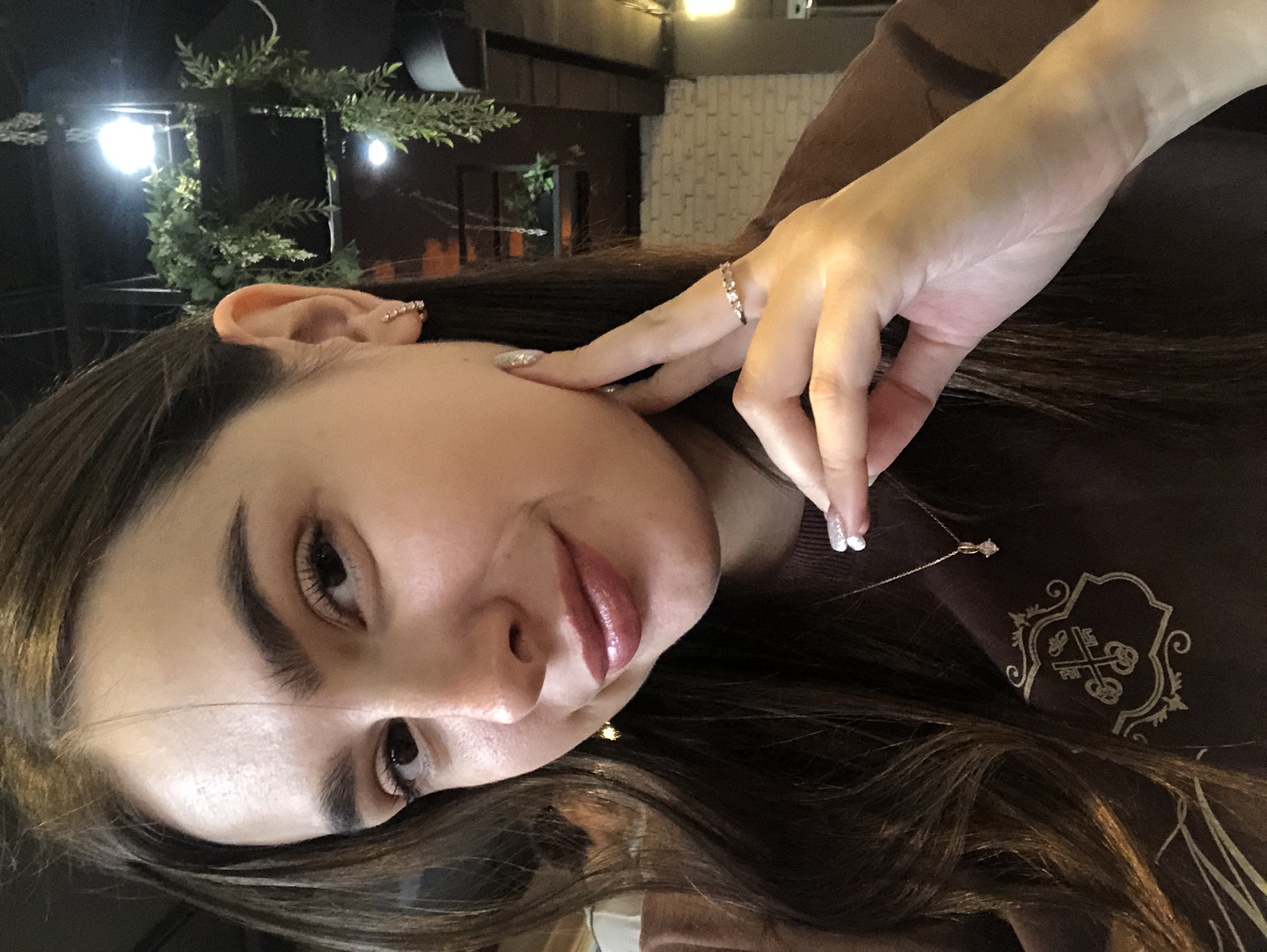 A young woman with long, dark brown hair is posing for a selfie. She is wearing a brown sweatshirt with a crest design on it and a delicate gold necklace with a diamond pendant. She has on a ring with small diamonds and a small hoop earring. Her left hand is gently touching her face, and her nails are painted with a glittery polish. The background appears to be an indoor setting, possibly a cafe or restaurant, with hanging plants and exposed brick walls. The lighting suggests an evening setting. There are no identifiable landmarks or contextual clues to pinpoint the location within Izhevsk, Russia.