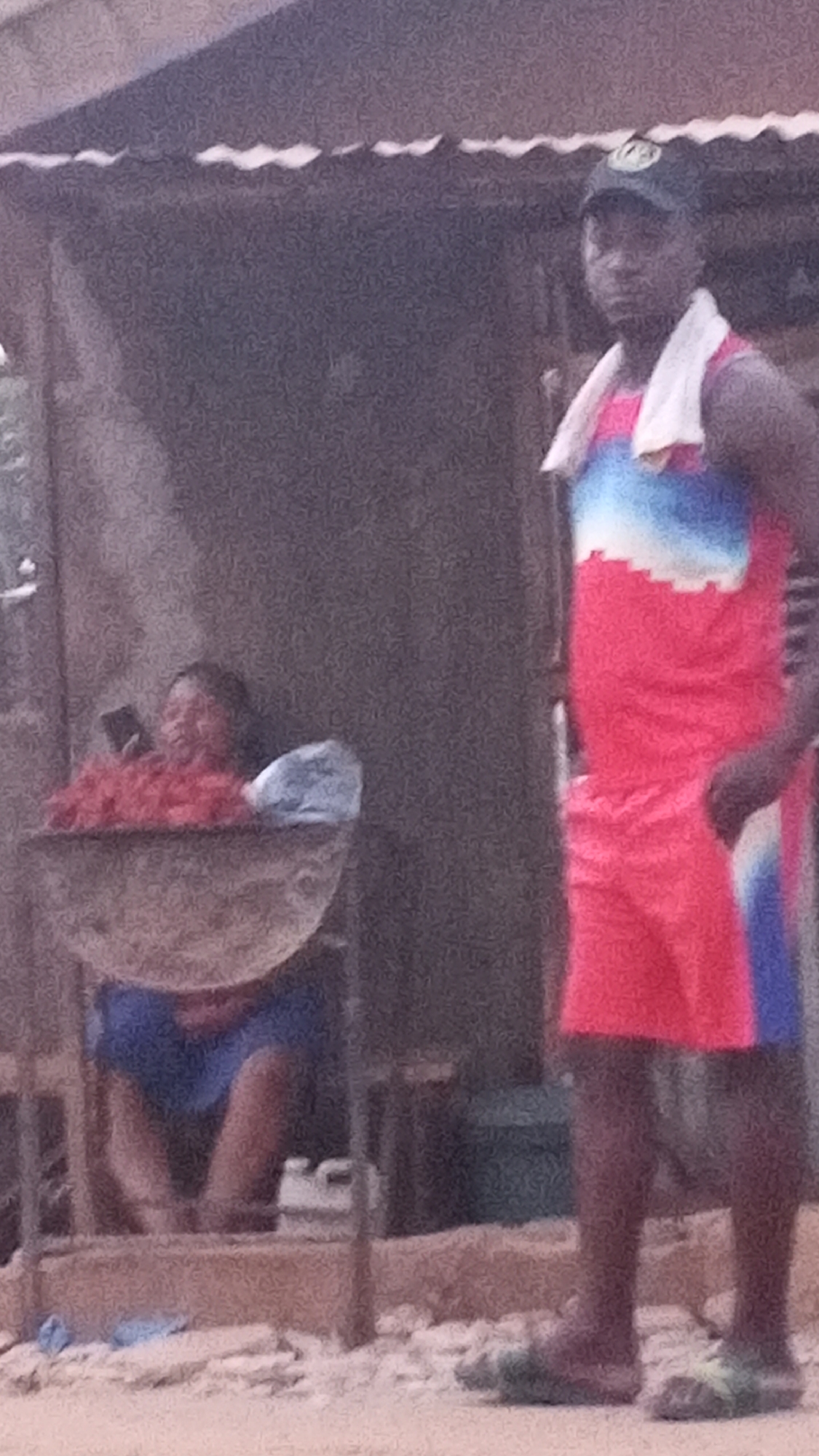 Sabon Tasha, Nigeria: A man in a red and blue tracksuit and a baseball cap stands barefoot in front of a corrugated iron structure, a white towel around his neck. Behind him to the left, a woman sits holding a phone behind a large bowl of what appears to be red peppers or other food items. The ground is sandy and littered with small debris.