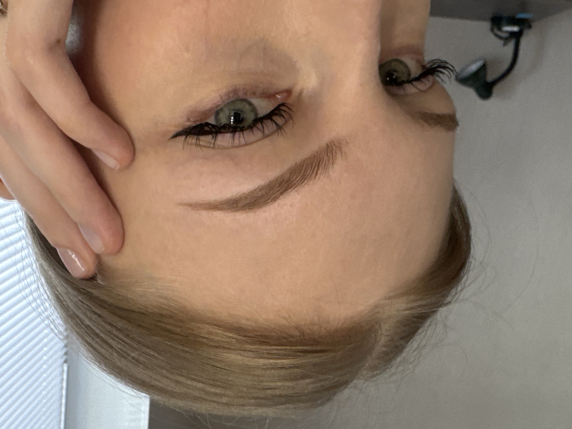 A close-up, rotated image shows a person's face. The person has fair skin, blonde hair, and green eyes. The left side of the face is partially covered by a hand. The person is wearing black eyeliner and false eyelashes. The background is a white wall with a black object (possibly a desk lamp) mounted on it. It is impossible to determine the location is Perm, Russia from this image.
