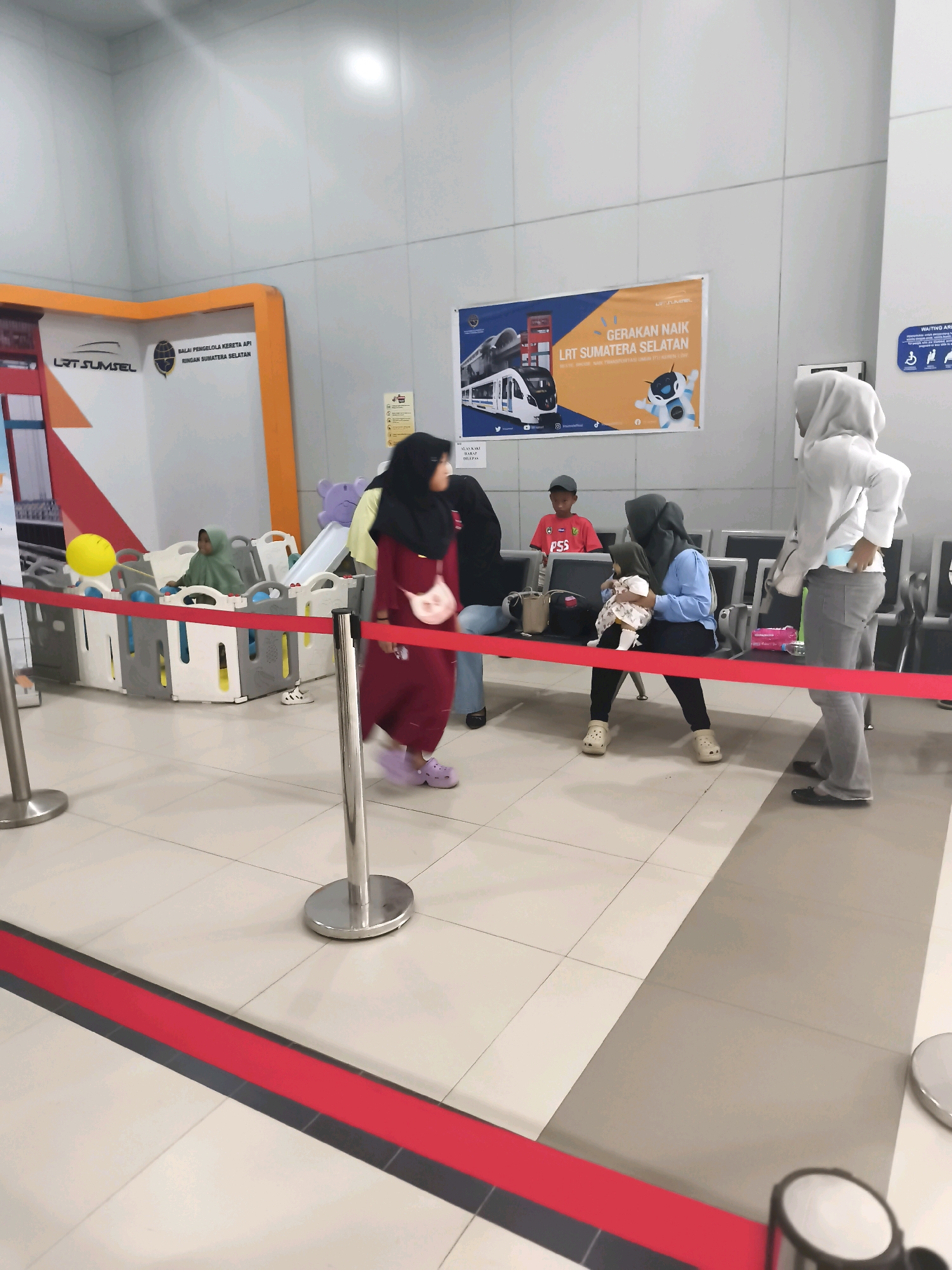 The image depicts an indoor waiting area, likely a station or terminal, in Kelurahan 16 Ilir, Kec. Ilir Timur I, Indonesia. A red rope barrier guides the flow of people. In the foreground, a metal stanchion stands to the left, and another is partially visible on the right.
Several individuals are present. A woman in a maroon dress and headscarf walks past the barrier towards the right, carrying a small pink purse. To her left, a young child in a green headscarf sits in a gated playpen containing a yellow ball and a slide. Further back, a boy wearing a red shirt with "PSS" printed on it stands.
A woman in a blue shirt and black pants is seated, holding a baby. Another woman, wearing a light grey hoodie and grey pants, stands to the right of the seated woman, facing away from the camera. The environment features light-colored tiled flooring and pale grey walls. A promotional banner featuring a train and text in Indonesian, "GERAKAN NAIK LRT SUMATERA SELATAN," is displayed on the wall. Chairs are arranged in rows behind the seated individuals. The lighting appears to be artificial, casting a bright glow from above.