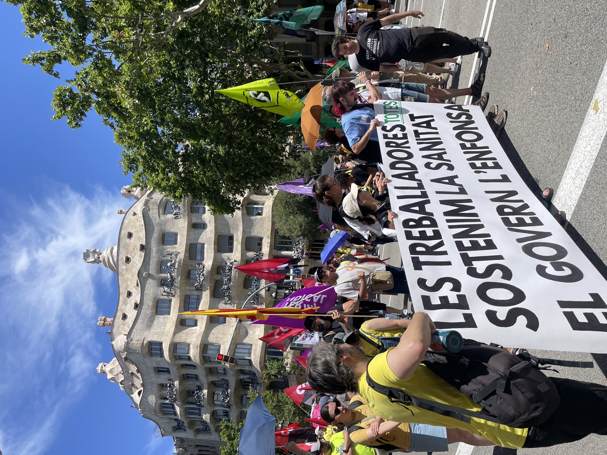 The image depicts a street protest in Barcelona, Spain, with a large banner carried by a group of people. The banner, held horizontally across the lower portion of the image, has large, bold black text that reads "LES TREBALLADORES TOTES SOSTENIM LA SANITAT EL GOVERN L'ENFONSA". Behind the banner, a crowd of protesters is visible, many holding flags of various colors, including red, yellow, purple, and green. In the background, a distinctive building with a wavy, organic facade and chimneys on the roof can be seen. This architectural style is characteristic of Antoni Gaudí's work in Barcelona. Green trees and a bright blue sky with some clouds are also present, suggesting an outdoor daytime event. People in the foreground are wearing casual clothing, and some are holding umbrellas or bags. The scene suggests a demonstration related to healthcare and government policy.