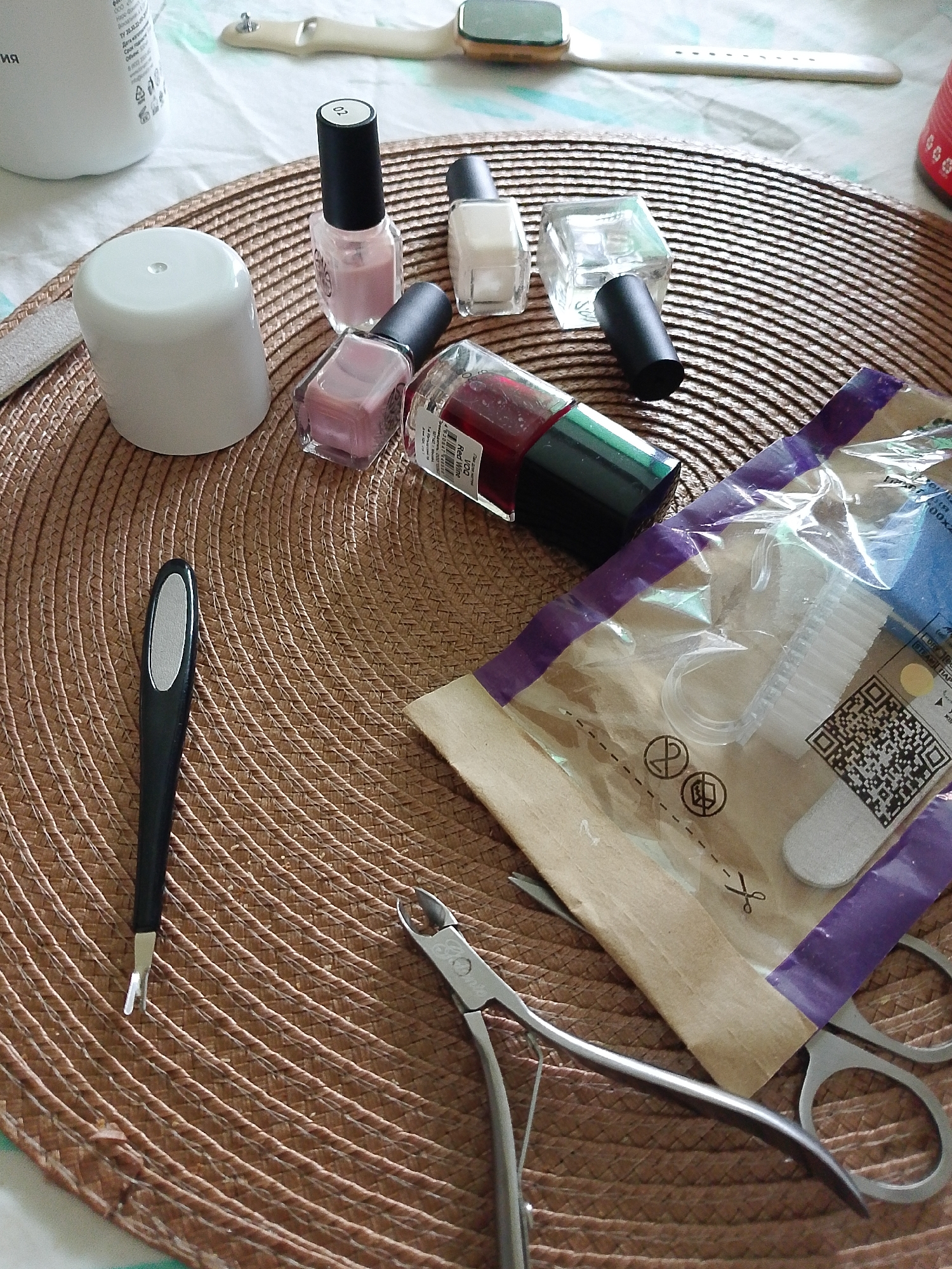 A round, woven placemat is the central feature of the image, upon which various manicure tools and supplies are arranged. These include several bottles of nail polish in shades of pink, red, and clear, a white bottle and container, and manicure implements such as nail clippers, a cuticle pusher, a small file, and a package containing manicure brushes and a small file. A white smartwatch with a light-colored band rests near the top of the frame, partially obscured. The background consists of a surface covered in white fabric with a subtle pattern. The items are generally clean and orderly. The location is not identifiable as Izhevsk, Russia based on the objects in this picture.
