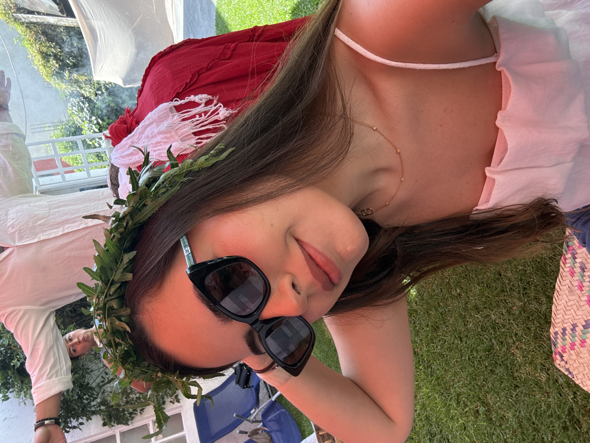 A woman wearing sunglasses and a leafy wreath rests on grass. She wears a light-colored sleeveless top and a gold necklace. A red garment with white fringe lies nearby. Behind her are white walls, green foliage, and a glimpse of blue chairs. Another person in a white shirt is partially visible in the background. The location is implied to be in the city of Puebla, Mexico.