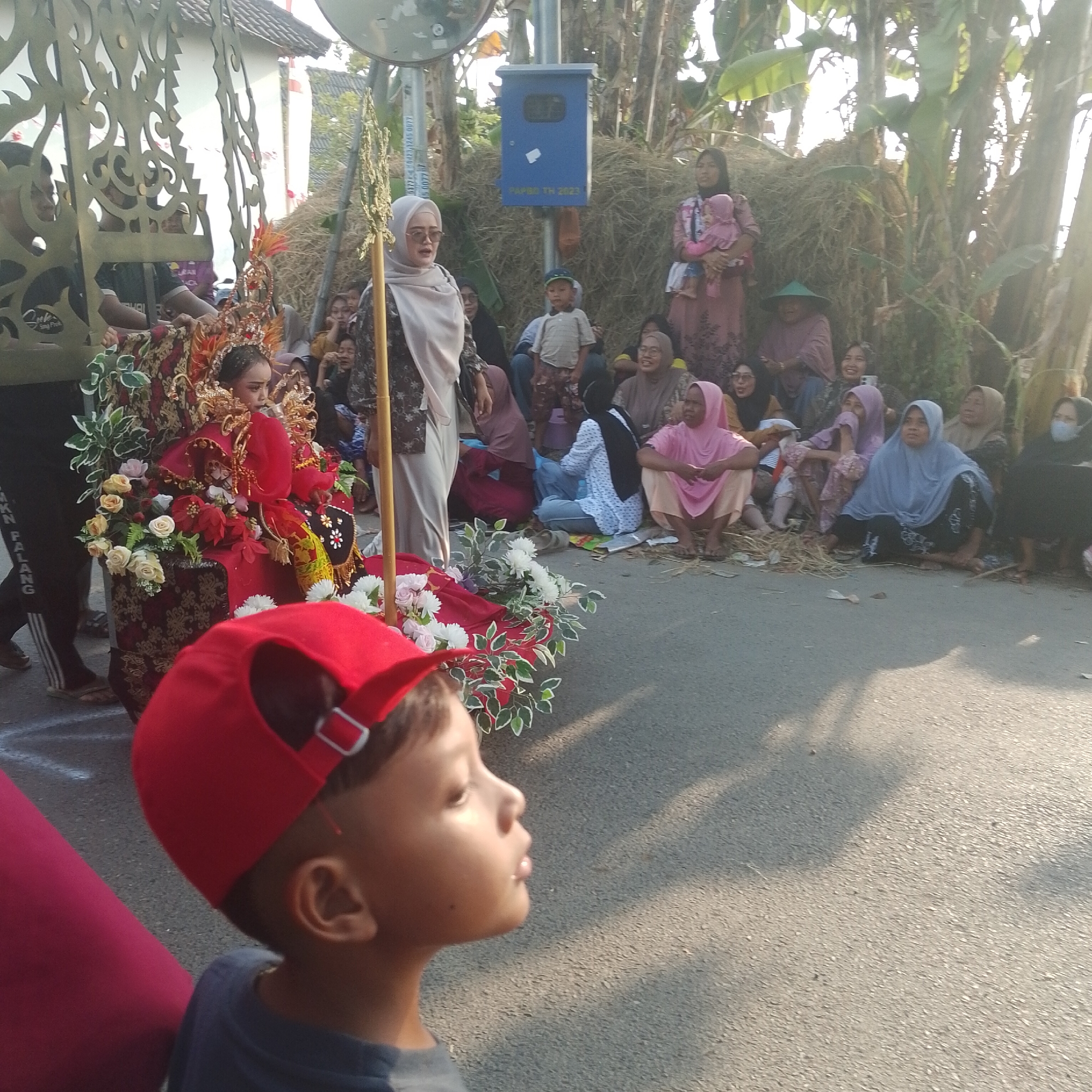 In Palang, Indonesia, a procession features a young girl adorned in a traditional red and gold outfit, complete with an elaborate headdress, seated on a decorated float. The float is embellished with red and white flowers and green foliage. Beside the float, people observe the event, some seated on the ground, others standing. A young boy in a red baseball cap is in the foreground, facing away from the camera. The background includes a haystack, trees, and buildings. A blue utility box is mounted on a pole. The scene suggests a cultural event or festival taking place outdoors on a paved road.