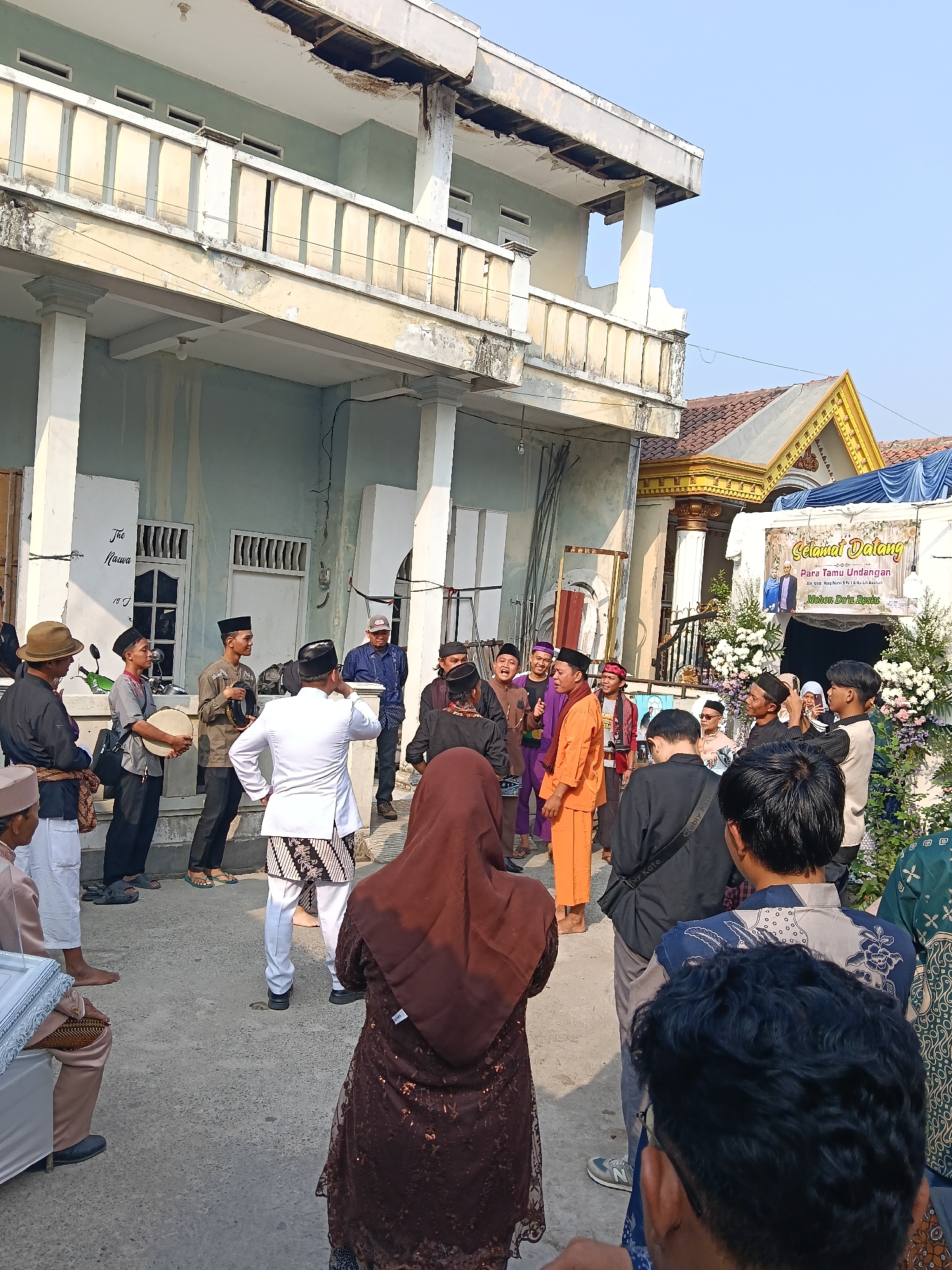 In Tegal Angus, Indonesia, a group of men and women are gathered outdoors in front of a two-story building. The building's facade is a faded green with white columns and a weathered balcony. To the right, a more ornate structure with golden accents and a red tile roof is visible, adorned with a banner reading "Selamat Datang Para Tamu Undangan" (Welcome Esteemed Guests) and floral decorations, suggesting a celebratory event like a wedding.
Several men are dressed in traditional Islamic attire, including white tunics, batik skirts, and peci hats. Some are holding musical instruments, possibly rebana drums, and appear to be participating in or preparing for a performance. Others are dressed in more casual wear. A woman in a brown hijab stands prominently in the foreground, facing away from the camera. The ground is paved with concrete. The scene is lit by daylight, with a clear blue sky overhead. The overall impression is of a community gathering for a significant occasion.