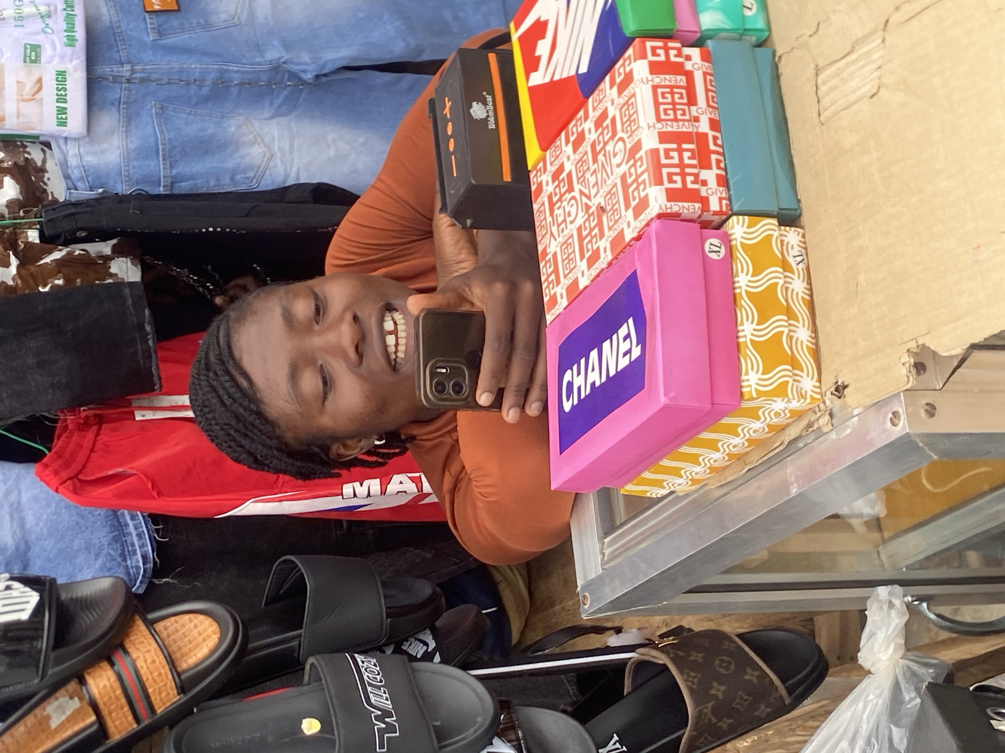 A young Black woman with braided hair, wearing an orange long-sleeved shirt, is looking at her smartphone in Bukuru, Nigeria. She is leaning on a display shelf filled with shoeboxes. The boxes are stacked in various colors and patterns, with visible branding such as "CHANEL" on a pink box and a red and white pattern on another. Behind her, several pairs of slides and clothing, including denim jeans and red shorts, are displayed on hangers. The scene appears to be an outdoor market stall, with a metal display case in the foreground.