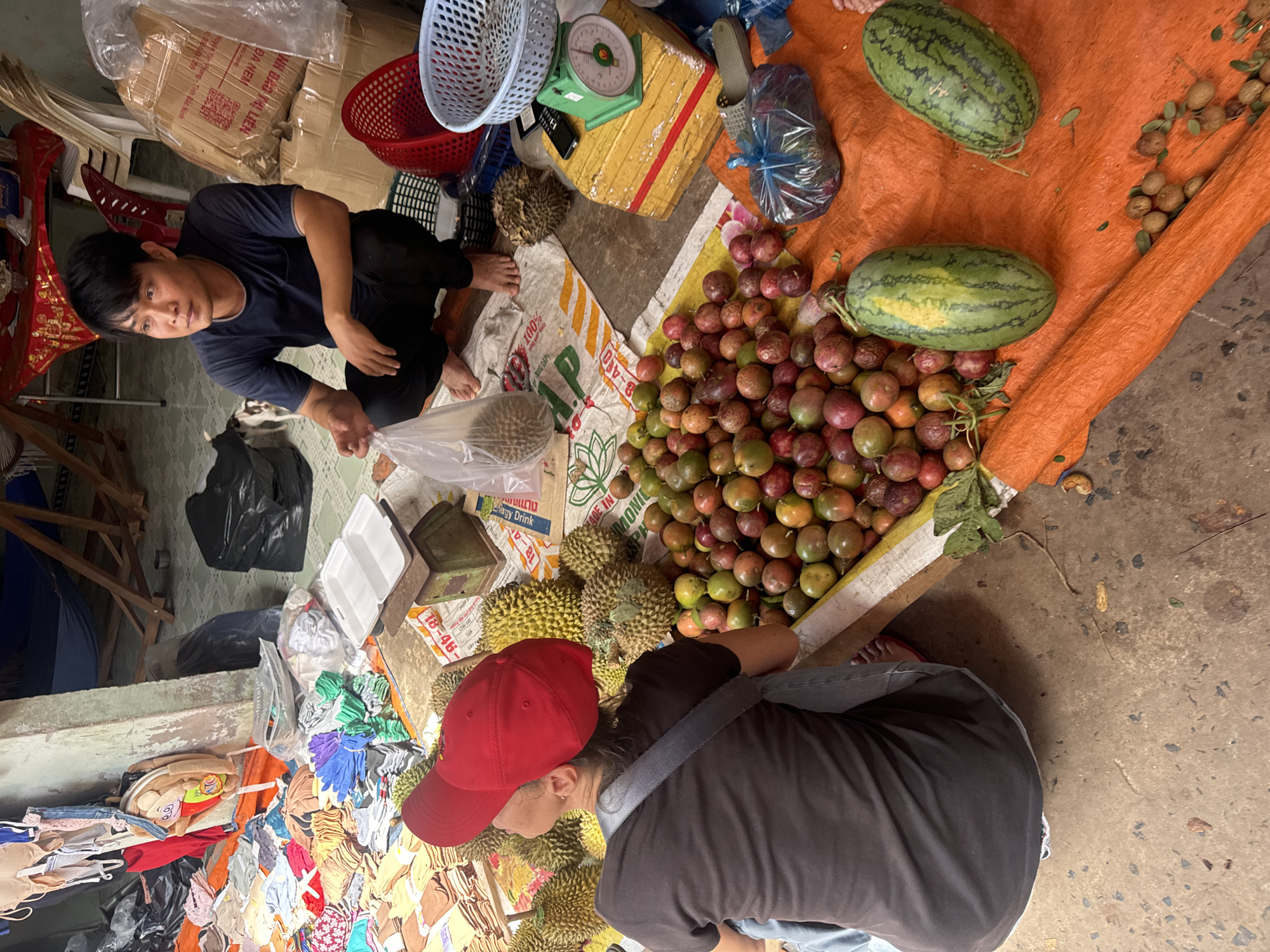 In Mũi Né, Vietnam, a street market scene unfolds. A man in a dark blue t-shirt and black pants crouches next to a pile of durian and other fruits, holding a plastic bag. Across from him, a person wearing a dark t-shirt and a red baseball cap is partially visible, facing away from the camera and looking at the fruit display. The ground is covered with a large orange tarp and a patterned cloth, on which various items are spread: durian, passion fruit, two watermelons, and smaller fruits resembling longans. In the background, a green weighing scale sits on a yellow box, and a red basket is to its left. More items, including clothing and black trash bags, are visible in the further background, suggesting a bustling market environment.