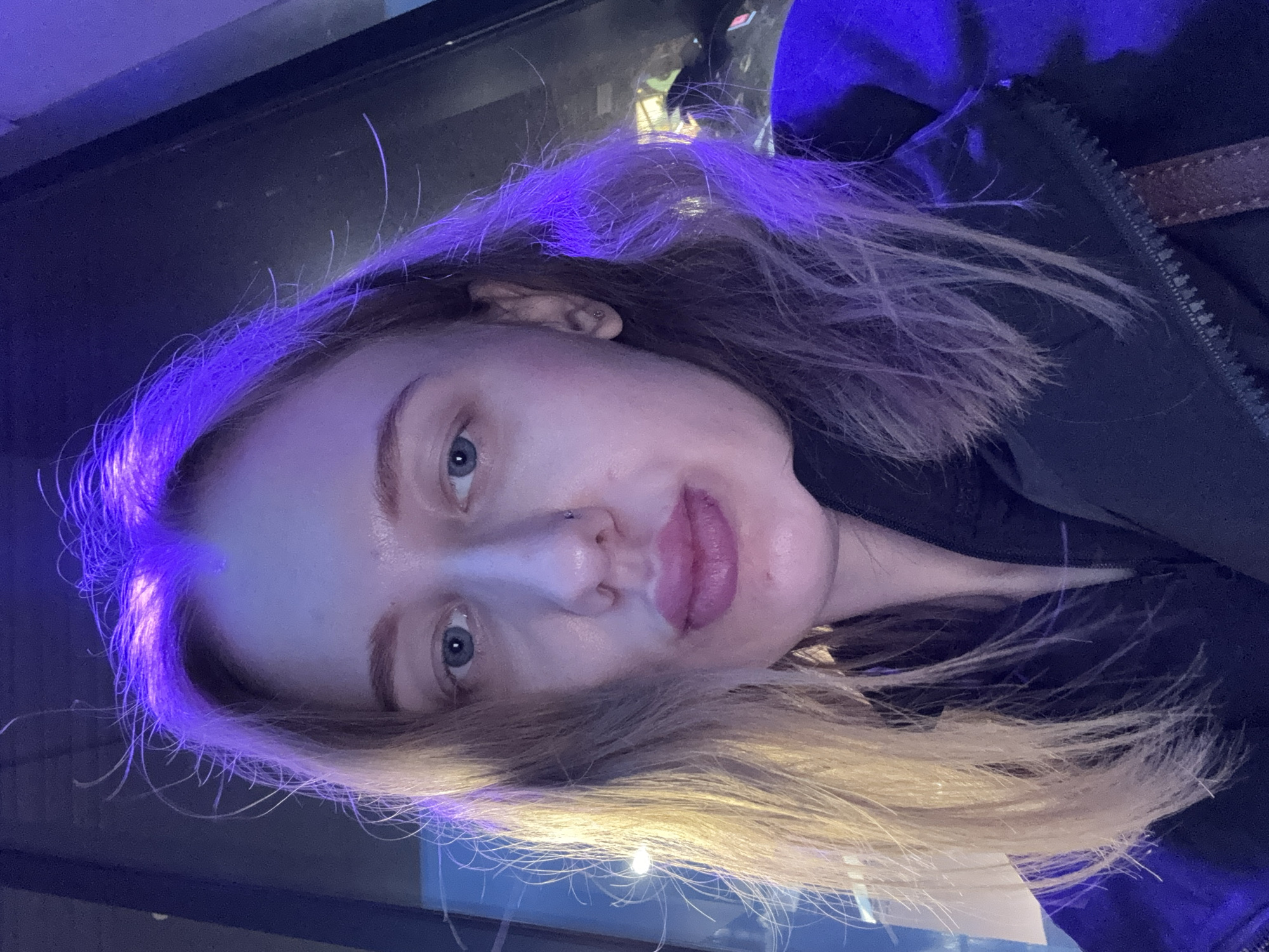 A selfie of a fair-skinned woman with blonde hair illuminated by purple lighting. The woman has blue eyes and a nose ring. She is wearing a dark jacket, possibly outdoors, in what appears to be an urban setting at night. The environment is dark with illuminated elements, possibly streetlights or other light sources reflected in a window or surface. There is no information about the location being Ol'ginskaya, Russia.