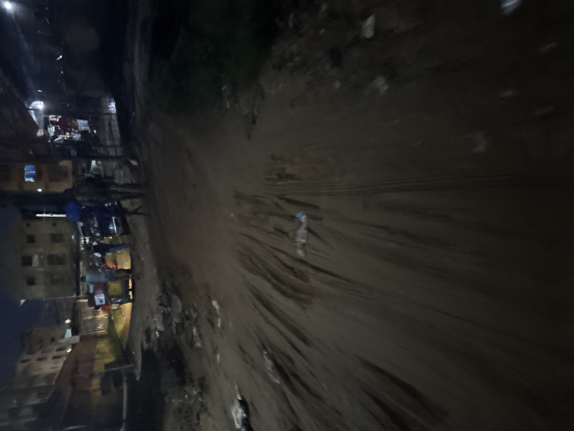 The image captures a dimly lit, unpaved street in Onitsha, Nigeria, at night. The ground is uneven and muddy, with visible tire tracks and scattered debris, including plastic bottles. In the mid-ground, a yellow tricycle, possibly a commercial taxi or personal vehicle, is parked. Several individuals are standing around the tricycle, some appearing to be passengers or vendors. Buildings with visible windows and varying architectural styles line the street, some illuminated, others dark. The overall atmosphere is gritty and utilitarian, characteristic of a busy urban environment at night.