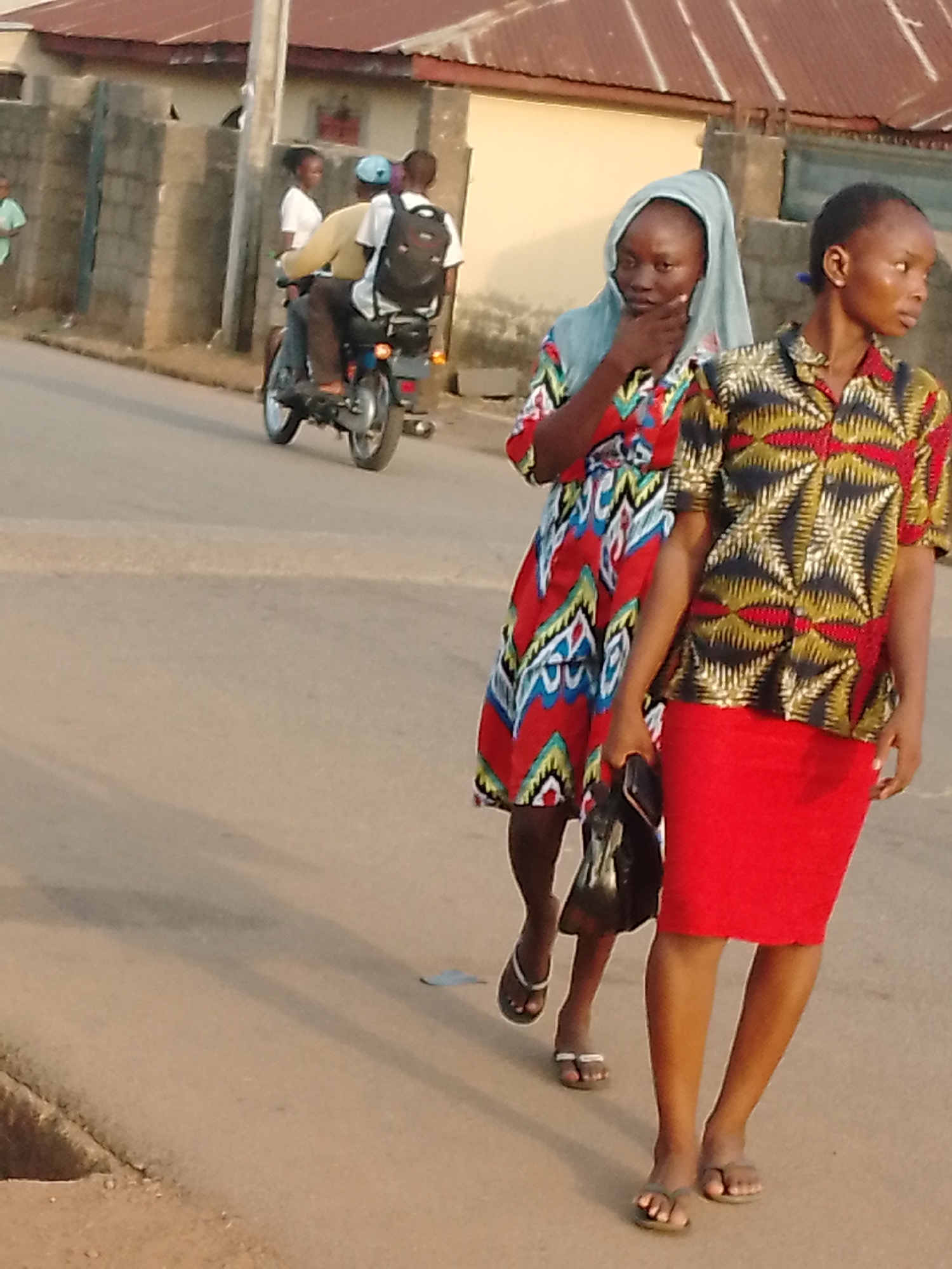 The image, taken in Akwanga, Nigeria, depicts two women walking on a street. One woman is wearing a red patterned dress and a light blue headscarf, with a black bag hanging on her left shoulder. The second woman is wearing a short-sleeved colorful patterned shirt and a red skirt. Both are wearing flip-flops. In the background, a man is driving a motorcycle with two passengers. The scene also includes a residential area with buildings and a fence. The overall impression is a street scene in a populated area with pedestrians and vehicular traffic.