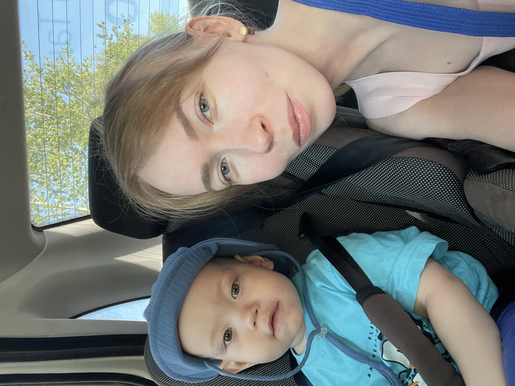 The image depicts a woman and an infant in a vehicle. The woman, who appears to be Caucasian, is fair-skinned with blonde hair and green eyes. She is wearing a pink top with a blue strap. The infant is wearing a light blue hooded shirt and is secured in a car seat with safety straps. The image appears to have been taken from inside the car, looking towards the back. The background shows trees and sky through the rear window. There is no information available in the image to confirm the location is in Perm, Russia.