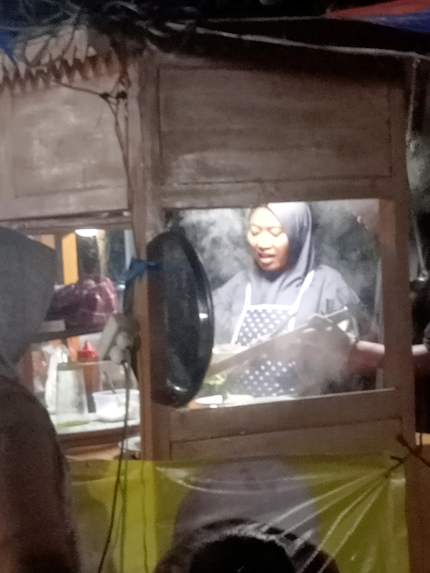 The image depicts a nighttime scene at a food stall in Tasikhargo, Indonesia. A woman wearing a grey hijab and a white polka-dotted apron is visible through a wooden window, appearing to be cooking or preparing food. Steam rises around her, indicating activity. In the foreground, parts of another person, also wearing a hooded garment, are visible, along with a yellow plastic covering that drapes over the stall. The overall lighting suggests an outdoor, possibly street food setting.