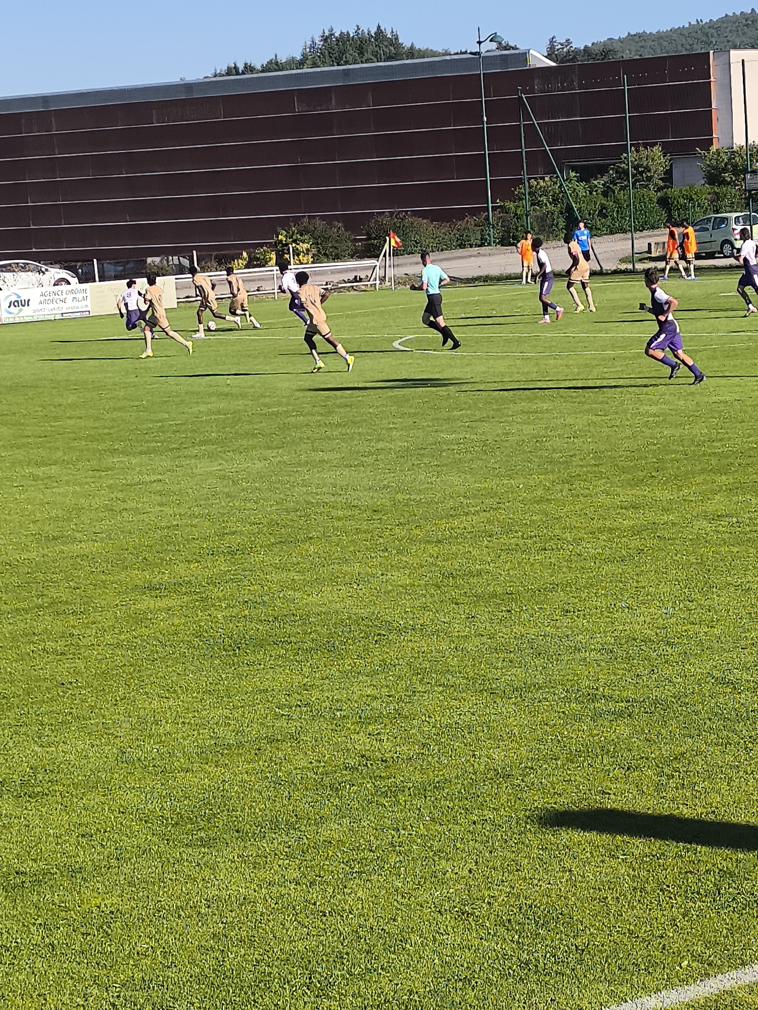 The image shows a soccer game in progress. Players wearing light brown and purple jerseys are running on a green field, while one referee is in a blue shirt and black shorts. There's a brown building in the background. The location is Maclas, France.