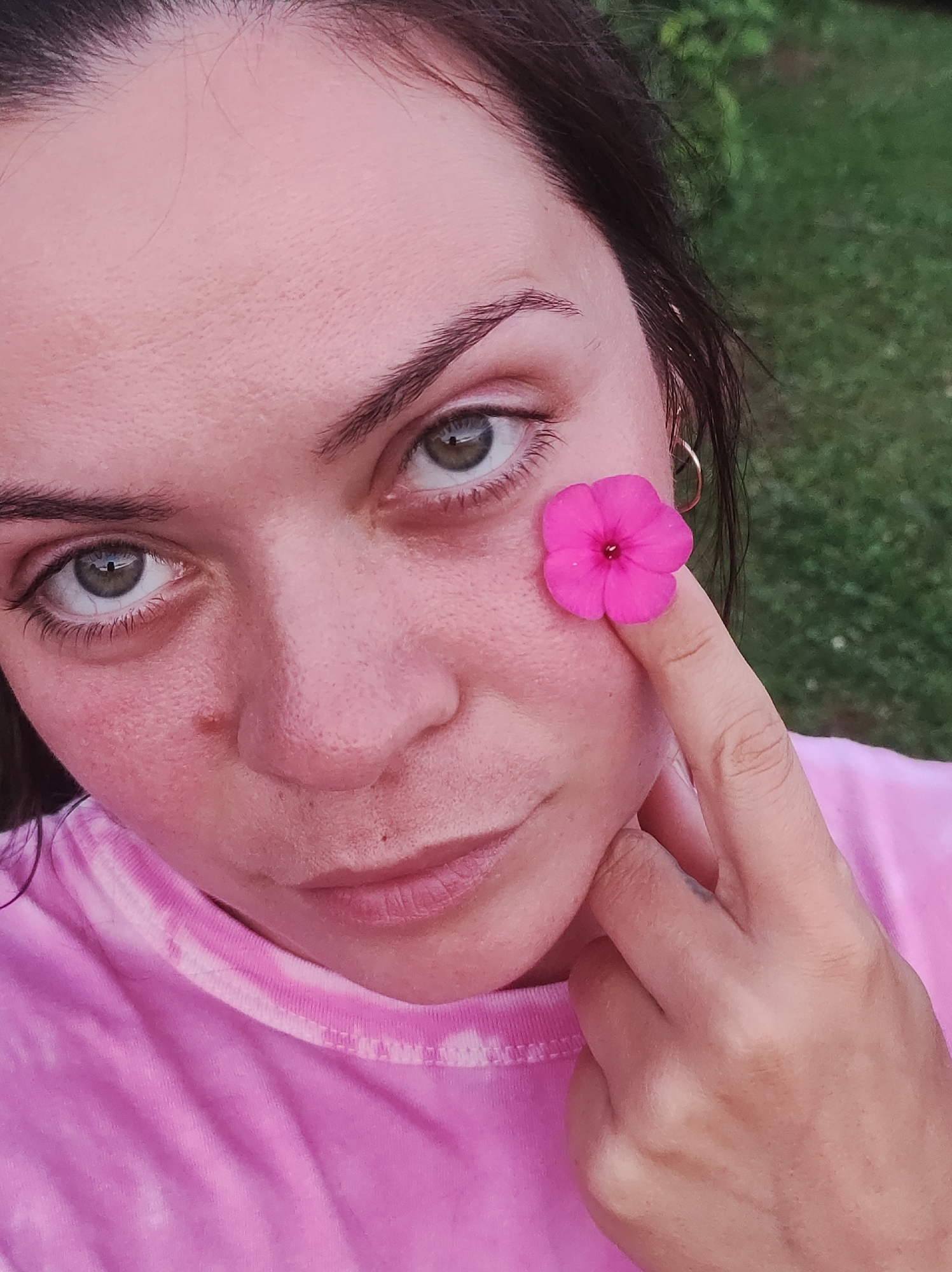 A close-up, low-angle shot captures the face of a young woman wearing a pink tie-dye t-shirt. She has dark hair and green eyes. A small pink flower is held against her right cheek by her index finger. Her left hand is resting near her chin, partially obscuring her mouth. The background is blurred green foliage, suggesting an outdoor setting. The woman's skin appears flushed, with some visible pores and a small mole on her right cheek. She is looking directly at the camera. The image composition focuses on her face and the flower, creating an intimate portrait. The location is not specified within the image.