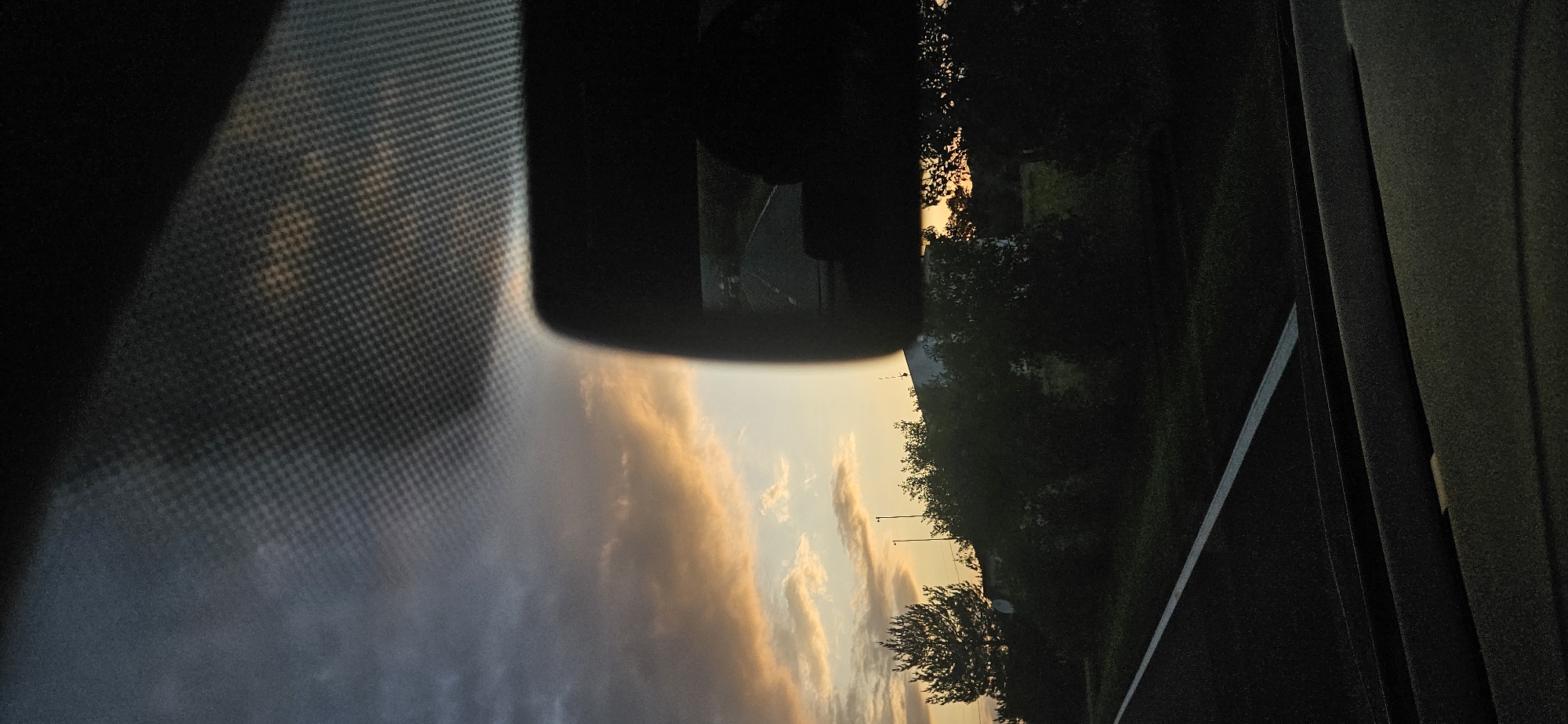 The image presents a perspective from inside a vehicle, likely a car, observing a twilight sky and rural landscape in Dolna Banya, Bulgaria. The upper portion of the windshield reveals a patterned frit band. A dark, rectangular rearview mirror is prominently positioned on the upper right, reflecting a segment of a road trailing the vehicle and an indistinct, dark shape, possibly a head or upper torso. The sky displays a gradient of colors, transitioning from darker blue-grey clouds in the upper left to warm orange and yellow hues nearer the horizon, indicative of either sunrise or sunset. Below the horizon, a dense line of dark green trees and bushes, including a distinct evergreen on the left, obscures much of the ground level. Faint outlines of structures or buildings are discernible behind the foliage. A road surface with a solid white lane marker is visible in the lower left quadrant of the frame. The vehicle's dashboard and interior elements are visible in the foreground, largely in shadow.