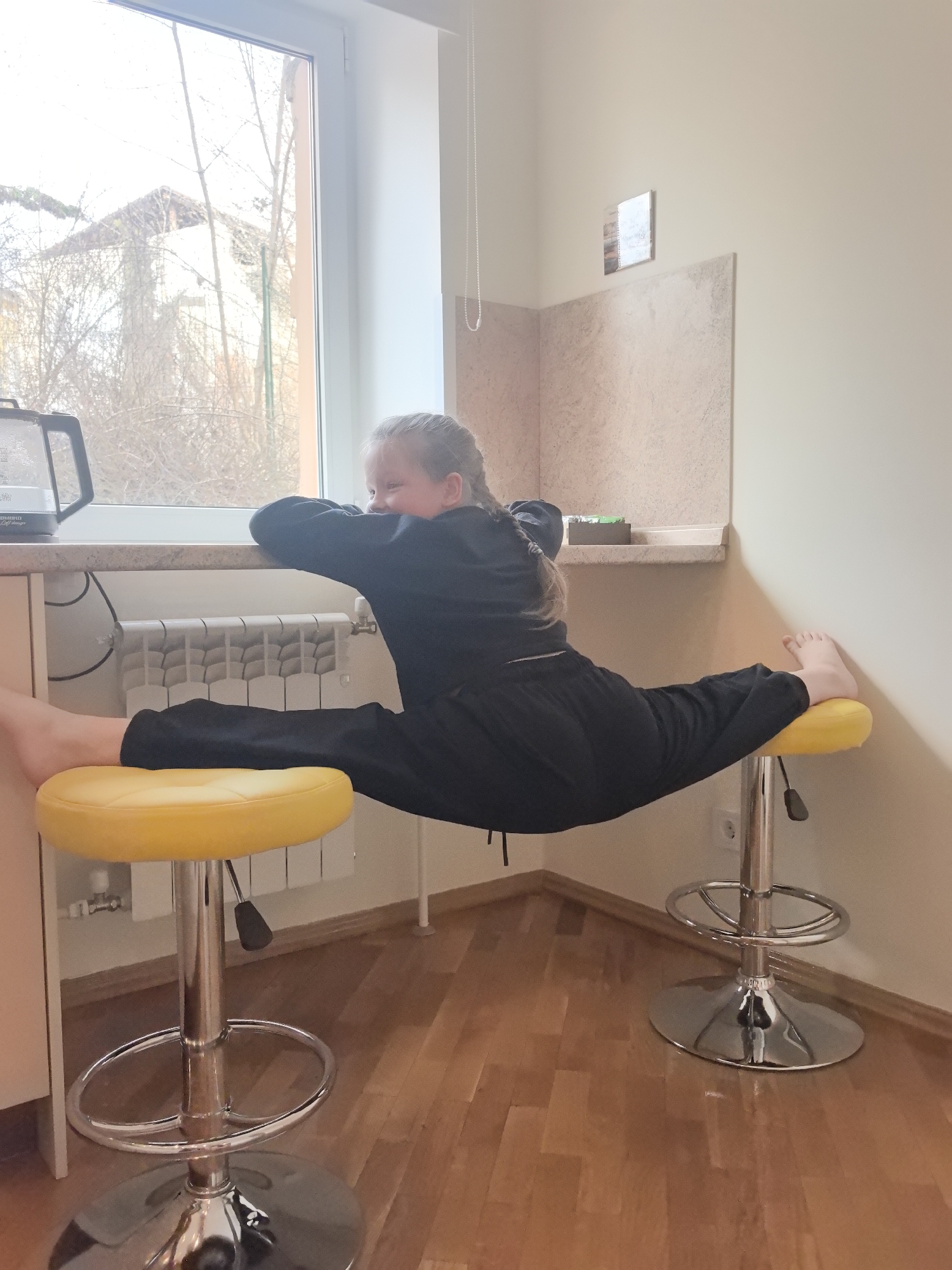 A young girl in a black tracksuit is doing a split between two yellow, chrome-based stools in a room with light beige walls and a light brown, wood-paneled floor. She is facing a window to the left, with her arms resting on the countertop. A radiator is visible beneath the countertop. A toaster is on the countertop next to the window, and there's a small shelf on the wall above the countertop. The location is in a room in a city of Nizhny Novgorod, Russia.