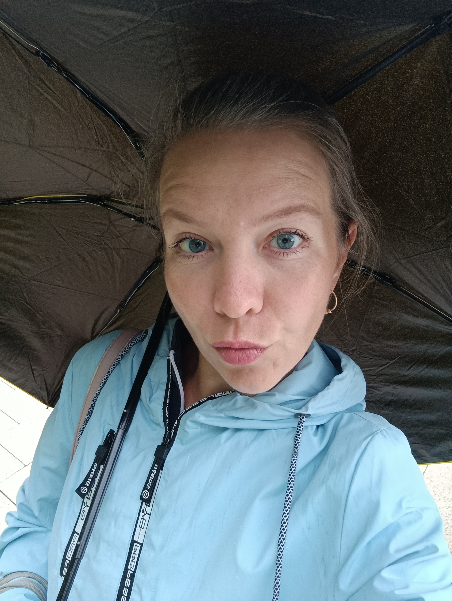 A woman is under an umbrella, possibly during a rainy day, taking a selfie. She has fair skin and light blue eyes, and is wearing a light blue hooded jacket with white drawstrings. She has a gold hoop earring in her left ear. The umbrella is dark in color, likely black or dark brown. The location may be the city of Chaikovsky, Russia, but no specific elements in the image confirm this.
