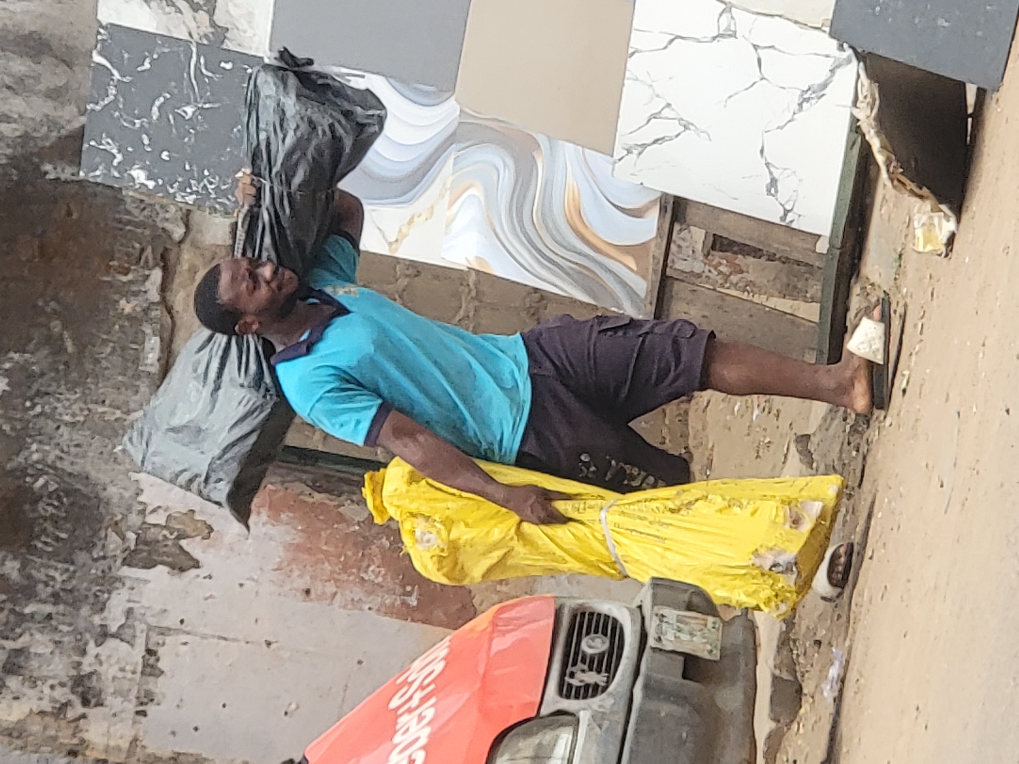 A man wearing a blue shirt and dark shorts carries two large bundles of goods in Maiburiji, Nigeria. The bundle on his shoulder is wrapped in dark gray material and the bundle in his arms is wrapped in bright yellow material. He is standing on a sidewalk next to a vehicle. Tiled display samples are visible behind him. The sidewalk and surrounding areas show signs of wear.