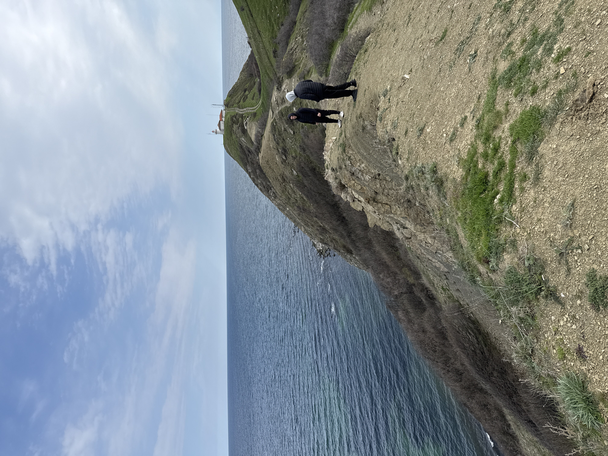 The image captures a coastal scene in Emona, Bulgaria, presenting a high-angle view of a cliffside overlooking the sea. Two individuals stand near the edge of the cliff, one wearing a white hoodie and dark clothing, and the other in dark attire. The cliff face exhibits a blend of exposed earth, rocks, and sparse green vegetation. In the distance, a building resembling a lighthouse or station is visible atop a grassy hill, connected by a path. The sky is partly cloudy, and the sea stretches to the horizon.