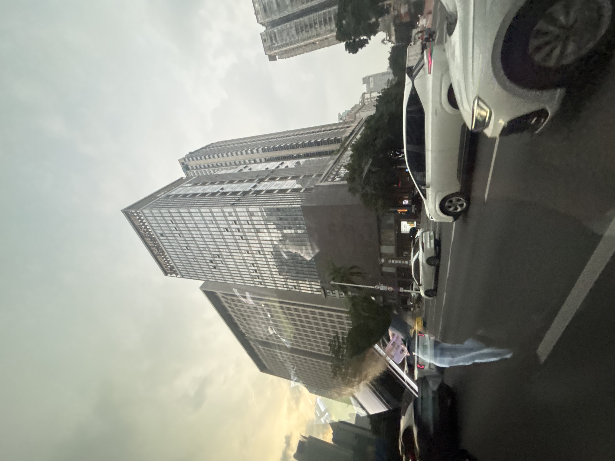 The image depicts a street scene in Tianhe District, China, characterized by tall, modern buildings with glass facades and a cloudy sky. Several cars are visible on the asphalt road, with some in motion and others stopped. In the foreground, a white car is prominently featured, its headlights illuminated. A blurred, translucent figure, possibly a person, is seen walking on the street, holding what appears to be a mobile phone. Trees line the street, and street signs are present. The overall atmosphere is urban, with a sense of a bustling city environment under overcast conditions.