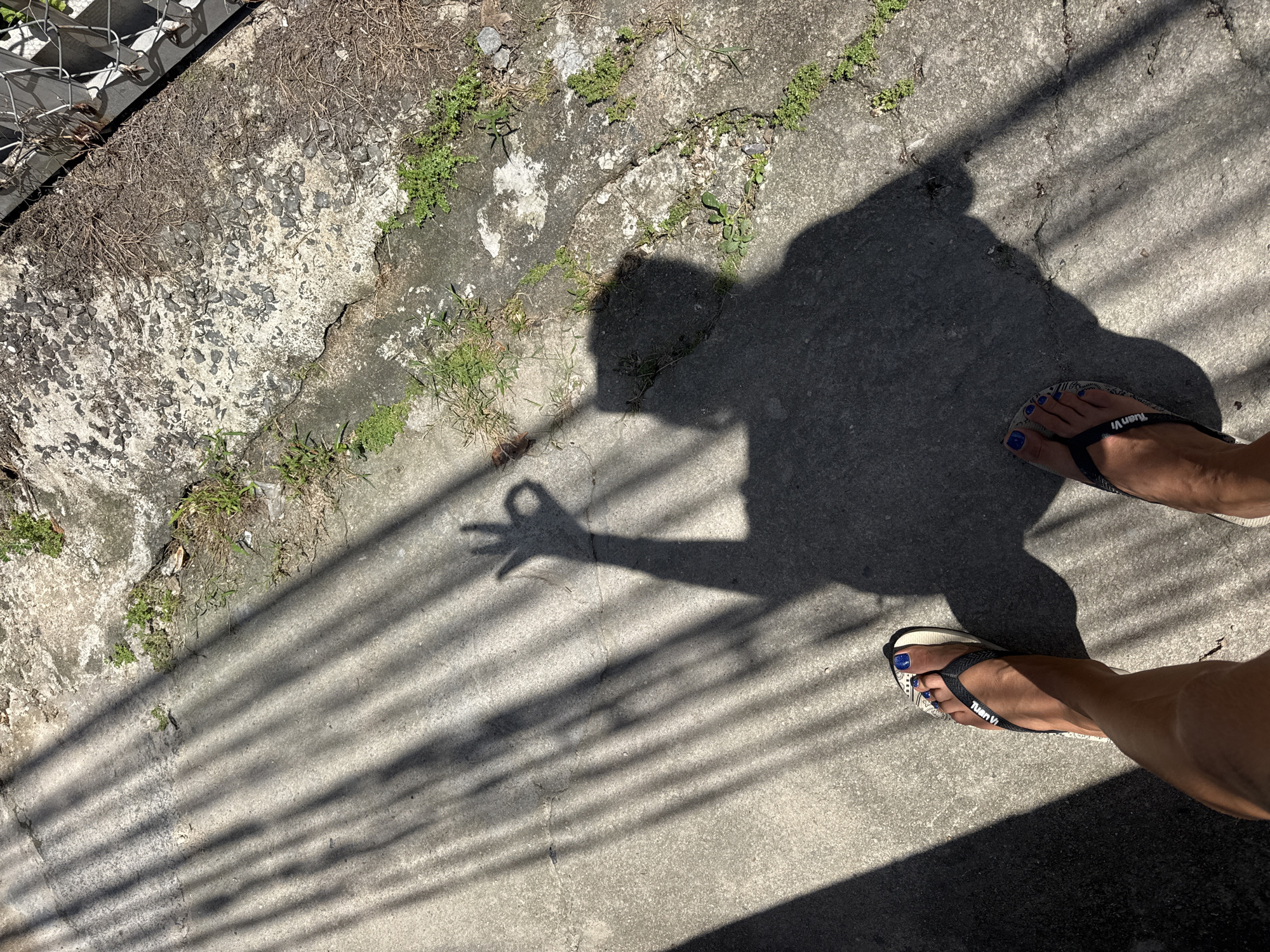 The image shows a shadow cast on a concrete surface, with vertical lines of light and shadow indicating a fence or similar structure. The shadow depicts a person's torso and head, with one arm raised, forming an "OK" hand gesture. The feet of the person casting the shadow are visible at the bottom of the frame, wearing flip-flops with a blue nail polish. The concrete surface is uneven and cracked, with some small plants growing in the cracks and along the edges. The overall scene suggests an outdoor environment in Bac Nha Trang Ward, Vietnam, likely a sidewalk or paved area.