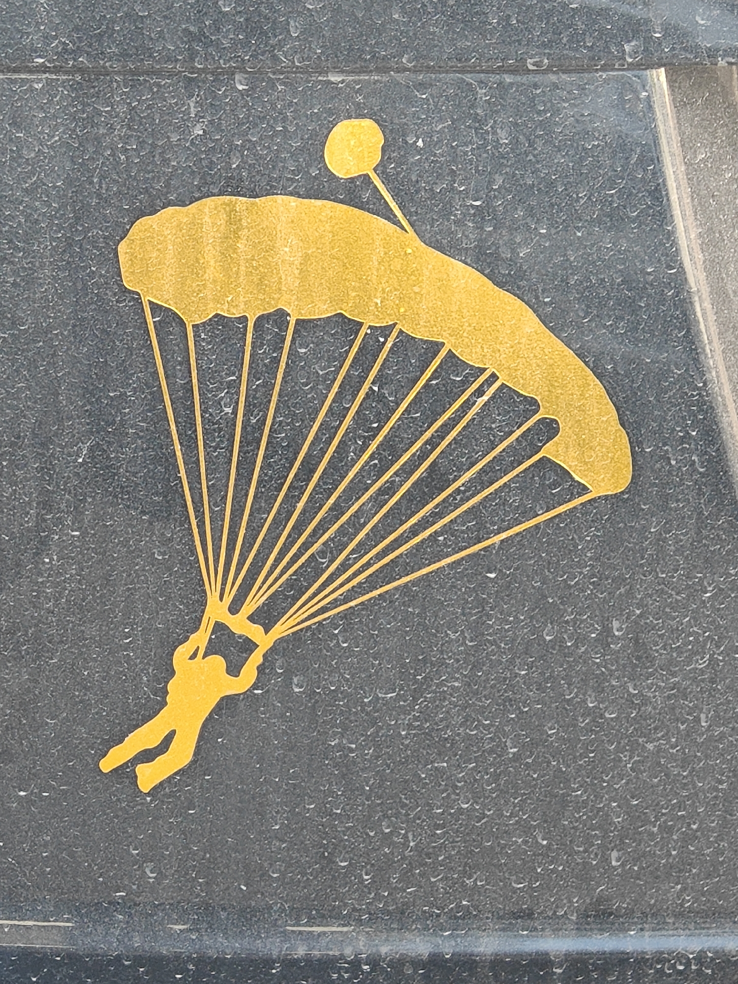 A yellow silhouette of a skydiver is depicted against a dark, speckled surface, possibly stone or metal, with small water droplets scattered across it. The skydiver is suspended mid-air, with arms outstretched, holding onto the lines of a large, open parachute. The parachute itself is a broad, curved shape, also in yellow, with numerous lines extending from its canopy to the skydiver. A small, circular yellow object, possibly part of the parachute rigging or a separate piece of equipment, is visible above and slightly to the left of the parachute. The overall impression is of a decal or emblem, highlighting the activity of skydiving. The lighting suggests it could be daytime, but no specific time can be determined. The presence of water droplets indicates it might be raining or has recently rained, and the surface's texture suggests an outdoor environment, though the specific location is unknown. There is no visible text in the image.
