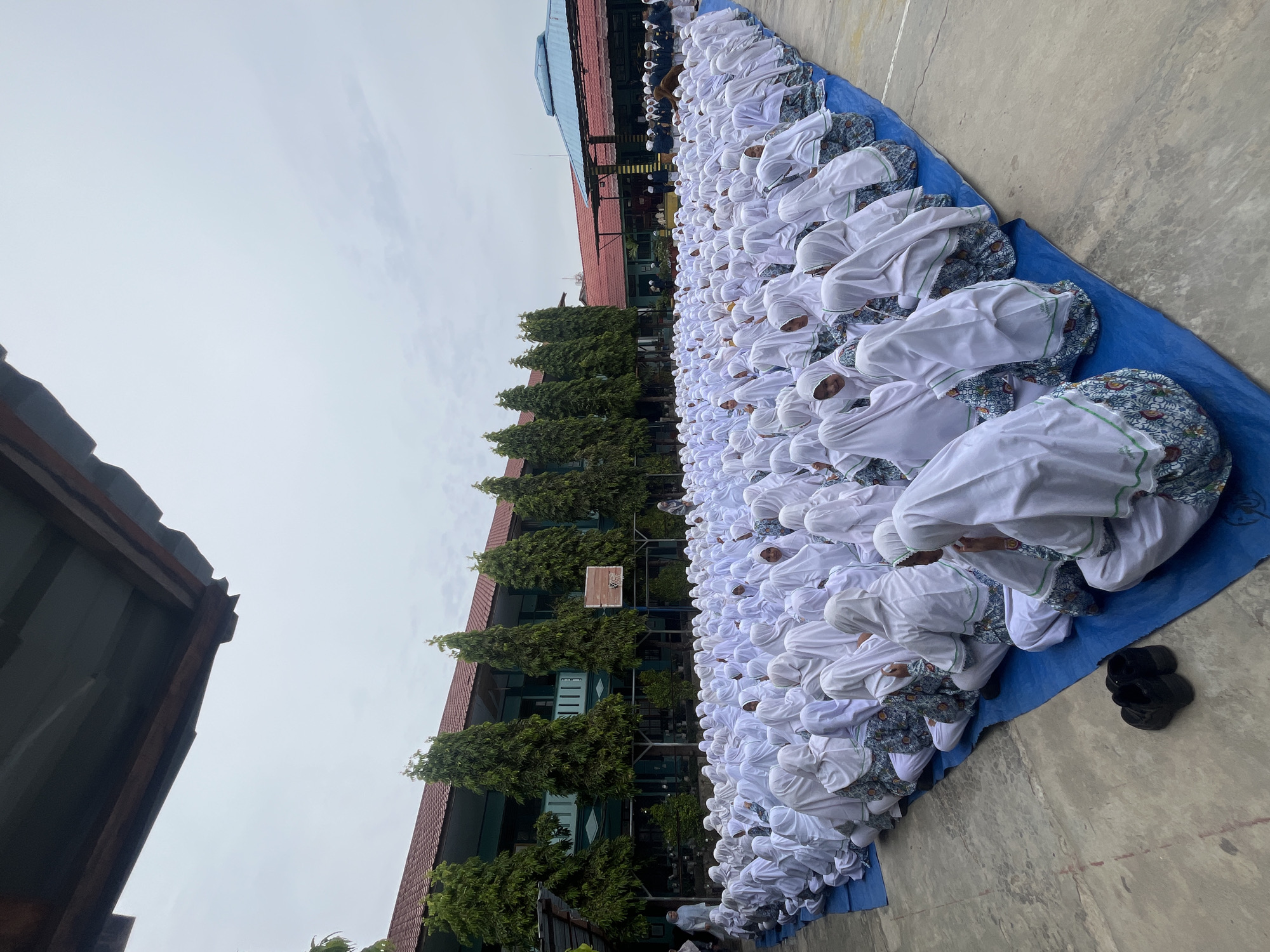 An outdoor gathering of an estimated 300 individuals, predominantly female, dressed in white hijabs and patterned skirts, is taking place on a blue tarp spread across a concrete surface. The individuals are seated in rows, facing towards the background. Behind them, a two-story building with a red-tiled roof and green facade stands, lined with tall, slender trees. In the distance, a covered structure with a blue roof is visible. The sky is overcast. The image appears to have been taken in the city of Lhokseumawe, Indonesia.