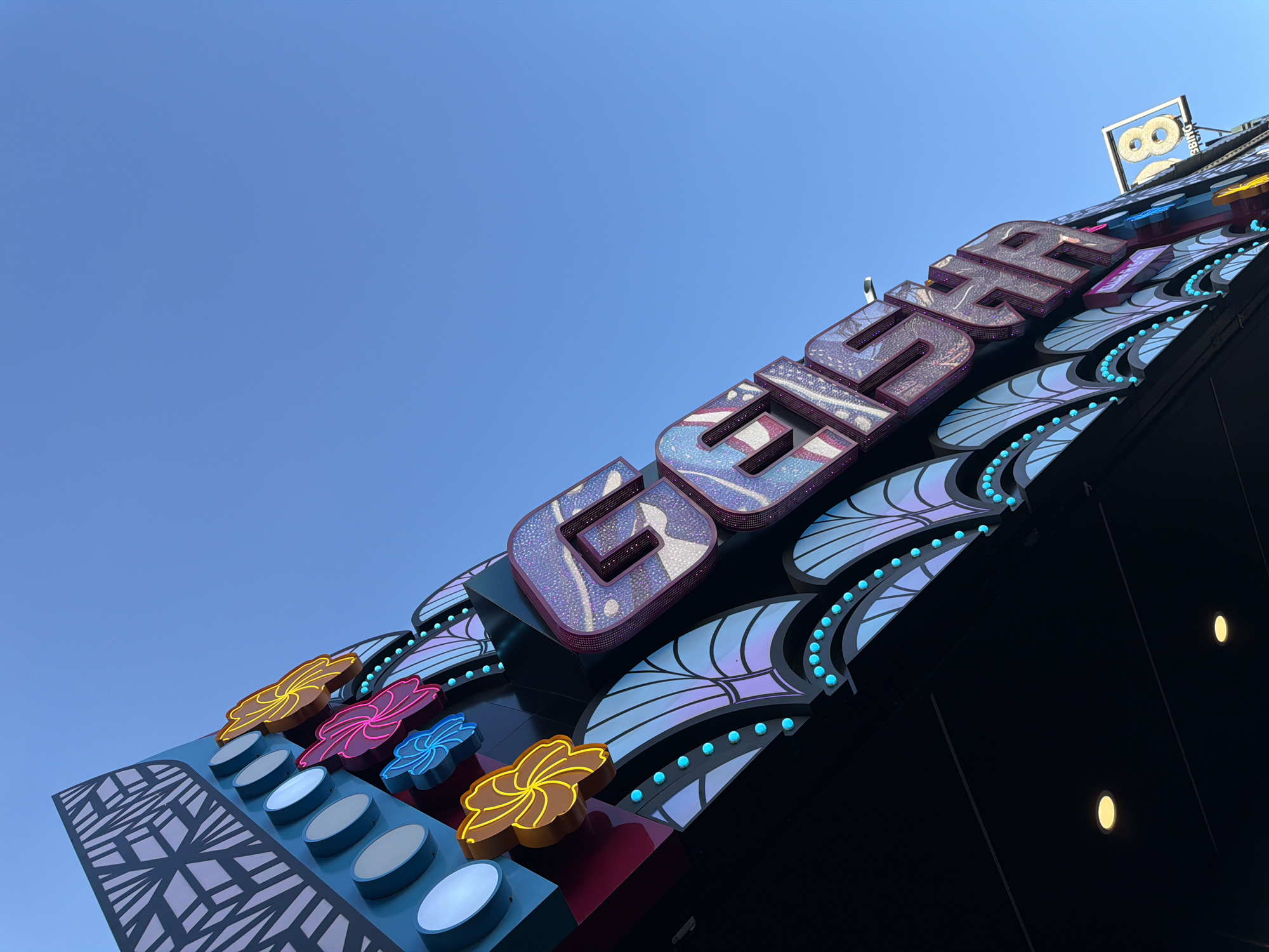 A low-angle, wide shot captures the brightly lit facade of a building against a clear blue sky in Pattaya City, Thailand. The building features large, stylized letters that spell out "BIG BUGSA" in a glittery, multi-colored design, with each letter outlined in a dark purple border. The letters are mounted on a dark, angled surface decorated with iridescent, fan-shaped patterns and rows of small, bright blue lights. To the left of the main sign are colorful, neon flower-shaped decorations and circular blue lights. In the upper right corner, a square sign displays a glowing "8" within a frame. The overall scene suggests a lively entertainment venue.