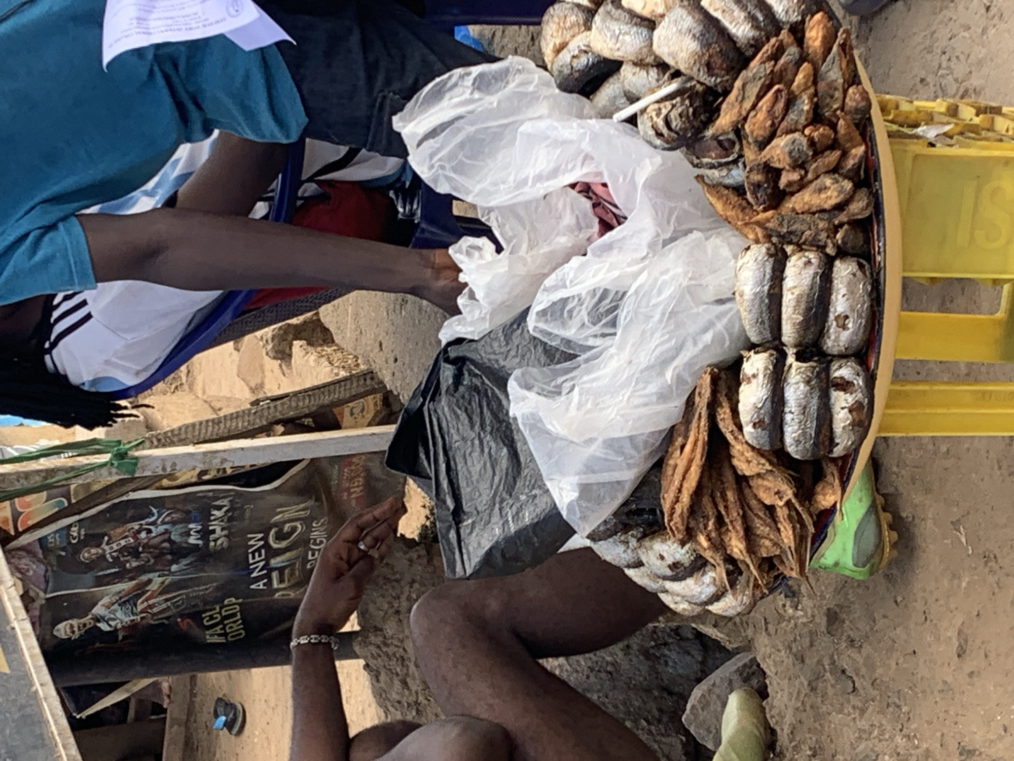 Captured from an elevated perspective, the image depicts a street-level scene in Rafin Par, Nigeria. Predominantly visible is an assortment of dried fish laid out for sale on a yellow plastic crate. The fish are of various sizes and types, some packaged in clear plastic bags. Two individuals are present: one seated on a blue plastic chair, clad in a blue t-shirt and black and white striped shorts, and another seated on the ground, wearing a silver bracelet. A crumpled poster is visible nearby, featuring images related to sports and entertainment. The surrounding environment consists of a rough, uneven ground surface.