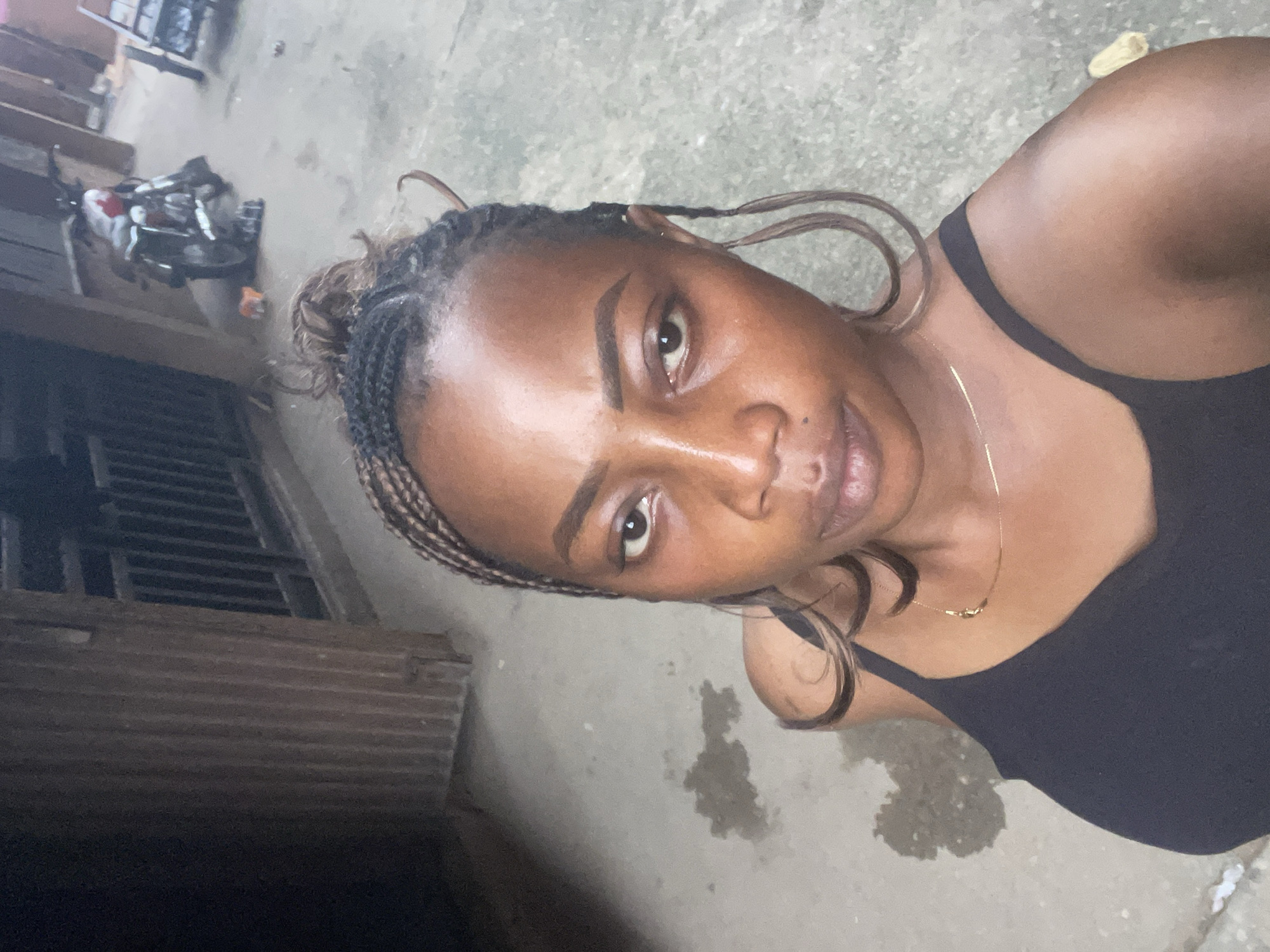A medium shot captures a young Black woman from the chest up, facing the camera. She has her hair braided in thin cornrows, pulled back into a bun, with a few strands loose around her face. She wears a black tank top and a thin gold necklace. Her left eye is open and looking towards the camera, while her right eye is partially closed, and her lips are slightly parted in a subtle smile. The background is a concrete ground with some damp patches. To the left, a corrugated metal wall with vertical ribs is visible, and to the right, a motorcycle and a cart are parked. The overall setting appears to be an outdoor urban environment, possibly a residential compound or a street in Fegge, Nigeria.