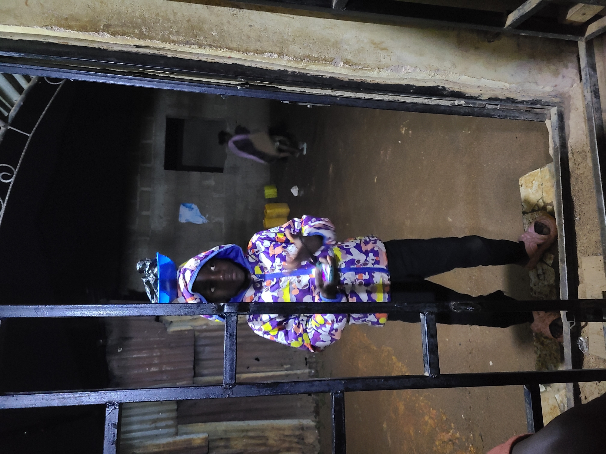 An individual, possibly a child or young adult, is standing directly in front of the camera, looking forward through a dark metal gate or grate. They are wearing a brightly colored, patterned hooded jacket with purple, white, yellow, and blue designs, black pants, and pink slip-on sandals. A small blue object with a silver lining rests on top of their head, and they hold a shiny object in their right hand.
The person is positioned slightly off-center, with the metal bars of the gate forming a grid across the lower half of the frame. Beyond the gate, the immediate area is a light brown, possibly dirt or concrete floor.
In the background, the setting appears to be a dimly lit, enclosed space. On the left, corrugated metal sheets are visible. On the right, block-built walls can be seen, with a dark, rectangular opening resembling a window or recess in one of the walls. Further back and slightly to the left, another person's torso and legs are visible, wearing what appears to be a light-colored top and possibly dark pants, moving away or standing still. Scattered on the floor in the background are a few objects, including some yellow items and a light blue plastic bag. The overall lighting is dim, suggesting it might be nighttime or a poorly lit indoor area. There is no visible text in the image.