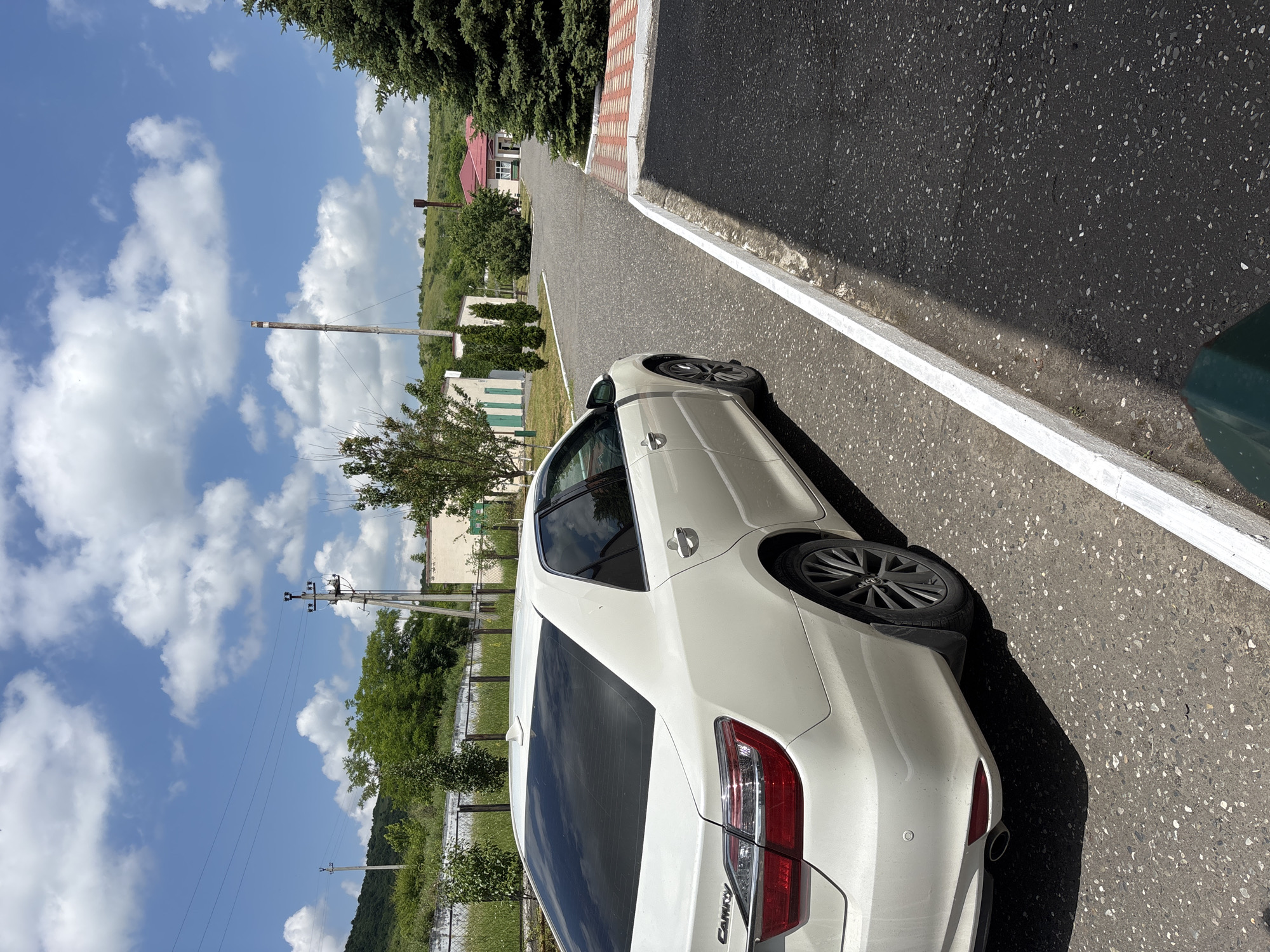 A white Toyota Camry is parked on an asphalt road. The car is facing away from the viewer and slightly to the right. The background shows a few buildings with light-colored walls and green accents, as well as some trees and rolling hills. The sky is blue with scattered white clouds. A tall, industrial-looking smokestack is visible in the distance. A large evergreen tree is on the right side of the frame. The road has a white concrete curb along the right side, separating it from a darker asphalt area.