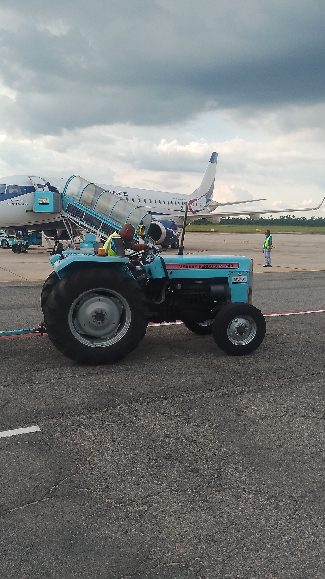 The scene is an airport tarmac in Orerokpe, Nigeria, under a cloudy sky. A blue "Massey Ferguson 240" tractor is positioned in the foreground. An airport worker is seated on the tractor, wearing a yellow safety vest. The tractor is towing a set of mobile boarding stairs, which are connected to an Embraer ERJ-145 aircraft of Air Peace, a Nigerian airline. A second airport worker, also in a yellow safety vest, stands further down the tarmac. The surface of the tarmac is cracked and shows signs of wear.