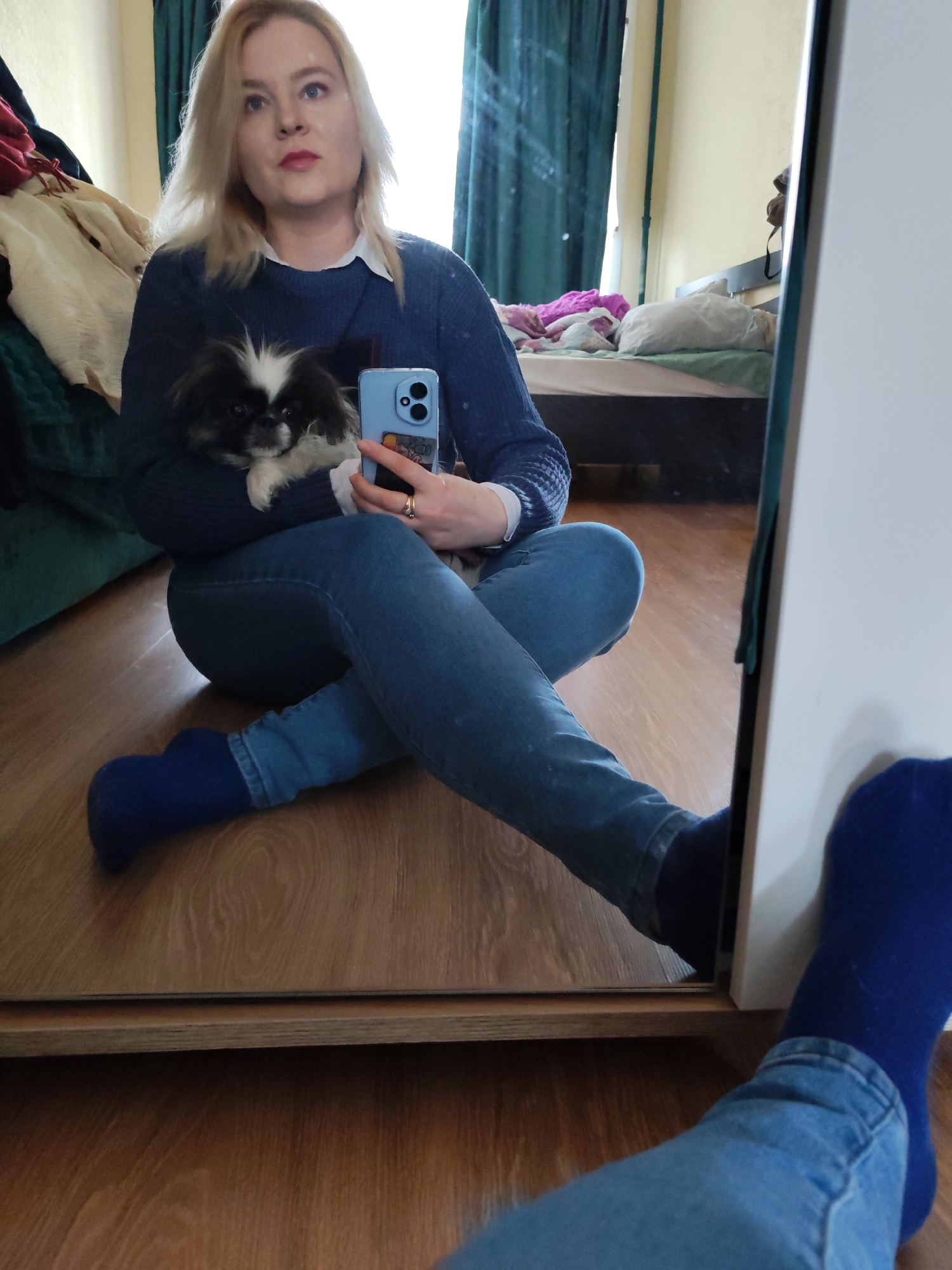 A woman with blonde hair is sitting cross-legged on a wooden floor, holding a small, black and white dog. She is wearing a blue sweater over a white collared shirt and blue jeans with blue socks. In her right hand, she holds a blue smartphone. The background includes a bed with a light green blanket and a dark green curtain. The image is reflected in a mirror, showing her legs and feet in blue socks. The scene is located indoors, likely in a bedroom. There are no definitive visual clues to place the location in Izhevsk, Russia.