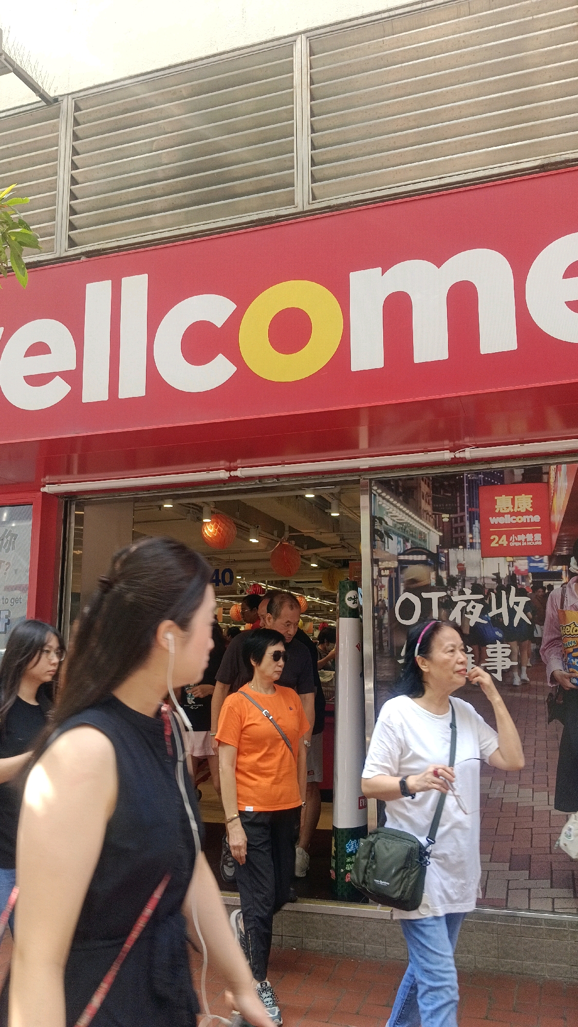 The image captures a street scene in Hong Kong, China, focusing on the entrance of a "wellcome" supermarket. The store's prominent red facade features large white lettering that spells out "wellcome," with the 'o' in the word depicted as a yellow circle. Above the signage, horizontal louvers are visible against a white building.
Several people are in the foreground and near the store entrance. In the left foreground, a woman with long dark hair, wearing a black sleeveless top, is partially visible. To her right, a young girl with long dark hair is also in view.
Moving towards the store, a woman in an orange t-shirt and black pants, wearing sunglasses, stands near the entrance. Behind her, a man in a dark t-shirt is also present. A woman in a white t-shirt and blue jeans, with a green shoulder bag, is walking towards the right side of the frame. Other individuals are seen inside the store and further down the street, suggesting a busy commercial area.
The entrance of the supermarket is an open doorway, revealing the interior with bright lighting and some display items, including orange spherical decorations hanging from the ceiling. Reflections on the glass doors of the supermarket show glimpses of the street and other buildings. A sign with Chinese characters and the word "wellcome" along with "24 小時營業" (24 hours business) is visible on the glass. The ground is paved with brick-like tiles. The overall environment appears to be an urban setting with commercial establishments.