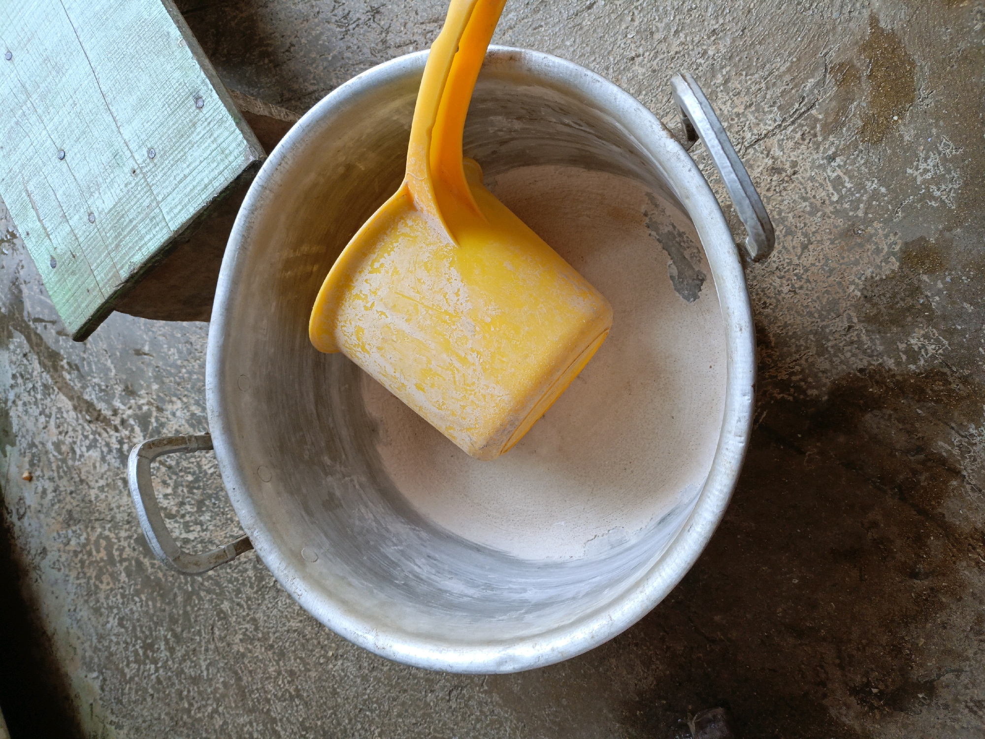 An overhead, medium-shot captures a close-up of a large, metal pot with two handles. Inside the pot, a yellow plastic scoop rests on a layer of light-brown powder. The pot is situated on a wet, concrete surface, stained with darker patches. To the left of the pot, a light-green wooden plank with several visible nails lies at an angle, appearing aged and worn. The overall scene suggests an outdoor setting, possibly in a yard or alleyway, with evidence of recent moisture on the ground. The image does not contain any discernible elements that would indicate its specific location within Jenu, Indonesia.