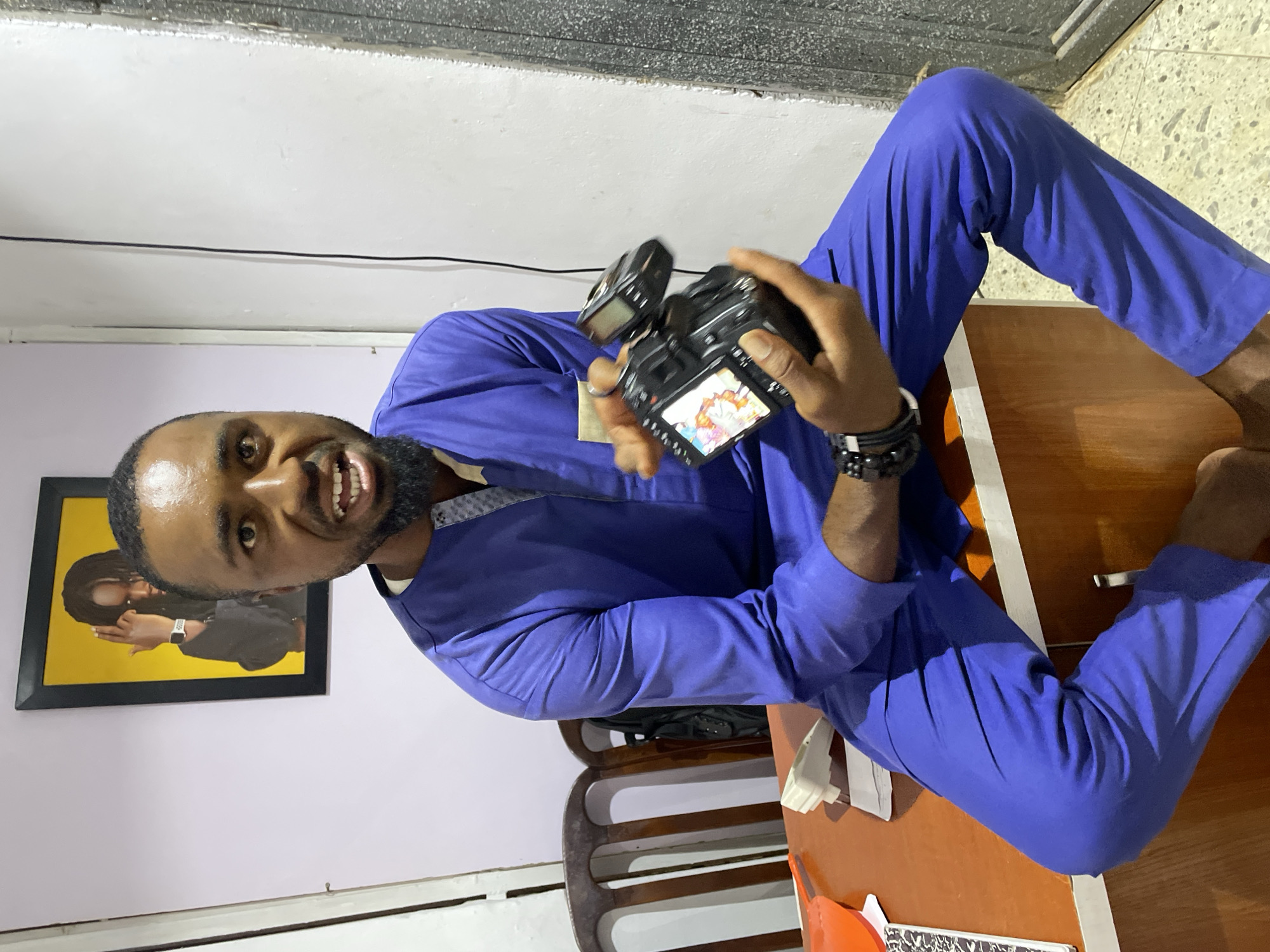 A man sits cross-legged on a wooden desk in a room. He is light-skinned and bearded, wearing a blue suit and dark beaded bracelets. He holds a black camera with a screen displaying an image. The camera's flash is raised. Behind him, a framed photo hangs on a light-colored wall. A wooden chair sits behind the desk. The floor appears tiled, and the ceiling is white with a textured border. The photograph was taken indoors, and based on the request it is likely the city of Jos, Nigeria.