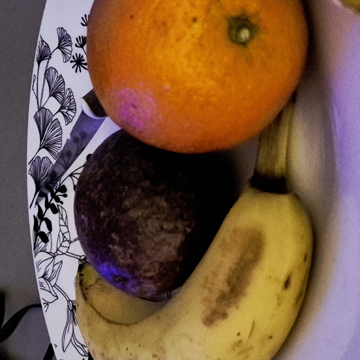 The image displays a close-up view of three fruits and a knife arranged on a white plate with a black botanical pattern. On the left, a yellow banana with several brown blemishes is positioned horizontally, curving slightly upwards. Adjacent to the banana, in the center, is a dark, rounded fruit with a textured, deep purple-brown surface, consistent with an avocado or a large plum. To the right, an orange citrus fruit is visible, featuring a small green dimple at its base. A silver-colored knife blade is partially visible, resting on the plate behind the dark fruit and extending towards the orange. The plate's design includes stylized black leaves, prominently featuring ginkgo-like motifs. The background is a blurred, plain grey, indicating an indoor setting. No individuals are present. The specific location within the city of Strasbourg, France, cannot be determined from the visual evidence provided in the image.