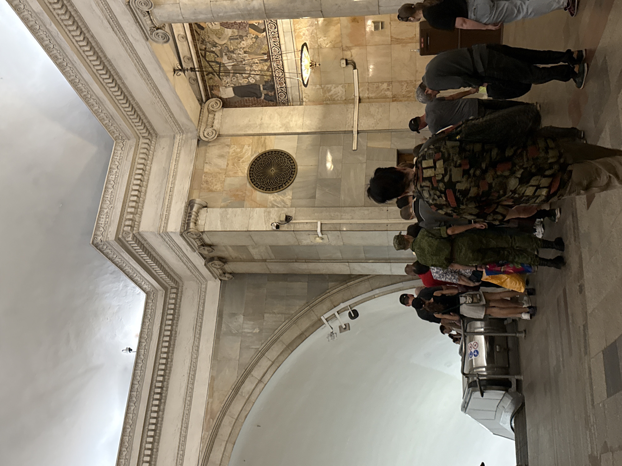 The image depicts a group of people queuing for an escalator in what appears to be a metro station in Moscow, Russia. The station's architecture is grand, featuring marble walls, ornate cornices, and a decorative mosaic mural above a large column. The ceiling is high and vaulted, with intricate molding. Several security cameras are visible, mounted on the walls and ceiling, indicating a monitored public space. The people in the queue are dressed in casual attire, with one individual in a military-style uniform. The escalator is at the lower left of the frame, leading down. The overall impression is of a busy, well-maintained public transport hub in an architecturally significant location.