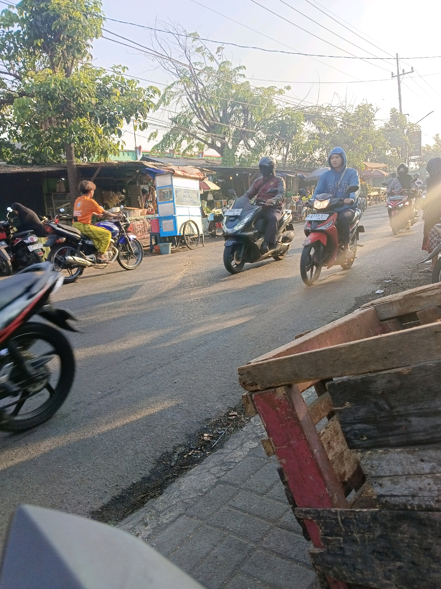 This image depicts a street scene in the city of Wonocolo, Indonesia, with several people riding motorcycles. In the foreground, a weathered wooden cart occupies the lower right portion of the frame. The road is paved with asphalt and lined with trees and small shops. Several individuals are on motorcycles, moving along the road. One person in an orange shirt and yellow pants is riding a blue motorcycle. Another individual wearing a blue hoodie is on a red motorcycle. Further down the road, other motorcycles and riders are visible. A light blue cart with wheels, likely a mobile vendor, is situated on the side of the road. The lighting suggests it is either early morning or late afternoon, with the sun casting long shadows. The environment appears to be an urban or semi-urban setting.