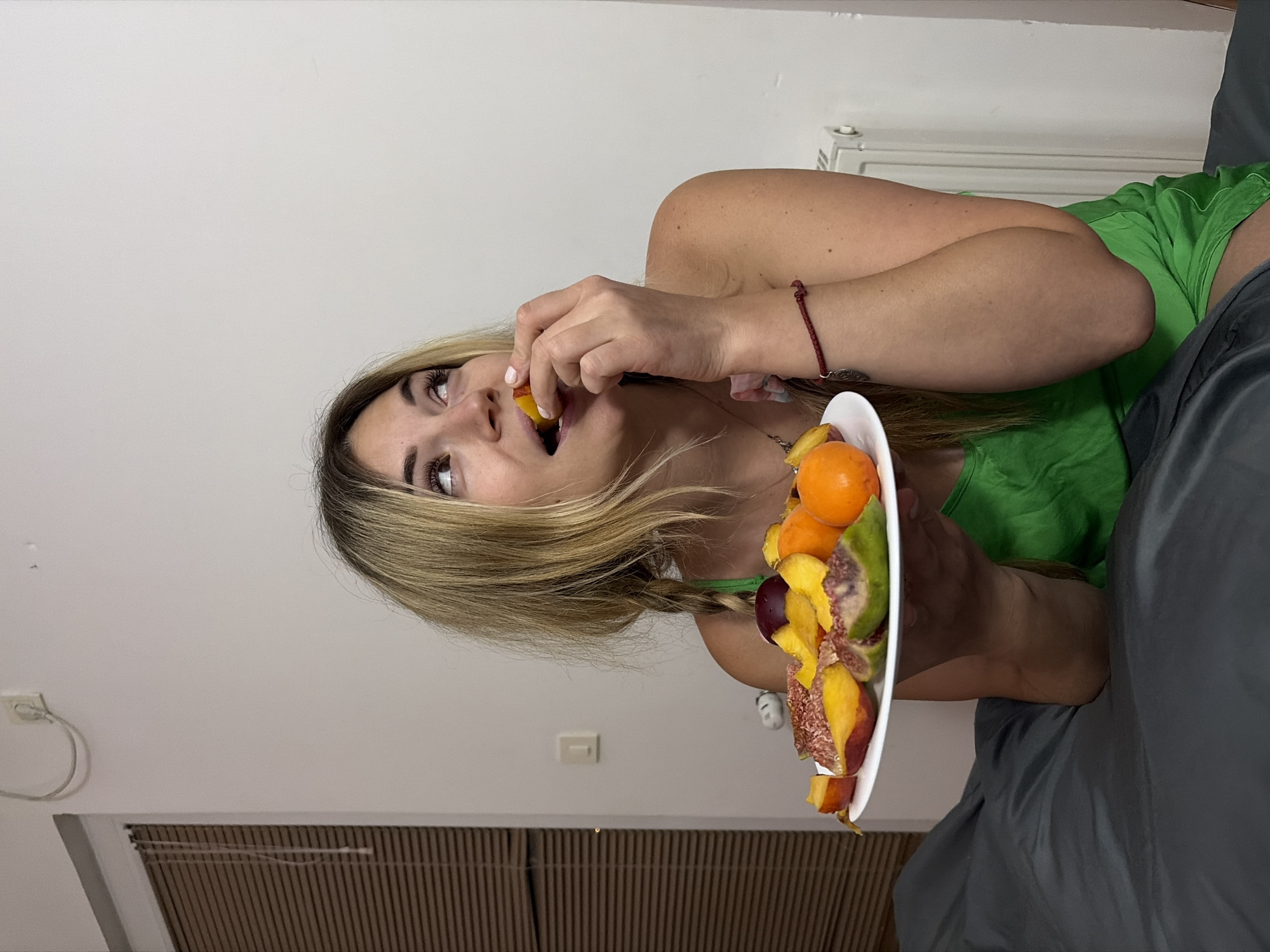 A young woman with blonde hair in two braids, wearing a green top, sits on a gray surface, holding a white plate filled with assorted sliced fruits, including peaches, figs, and tangerines. She is in the process of eating a slice of fruit, looking upwards and to the right. The background consists of a plain white wall with a light switch and an electrical outlet. To the left, a window with horizontal blinds is visible. A white radiator is present on the right side of the image. The overall setting appears to be a residential interior, possibly a bedroom, in Budva, Montenegro.