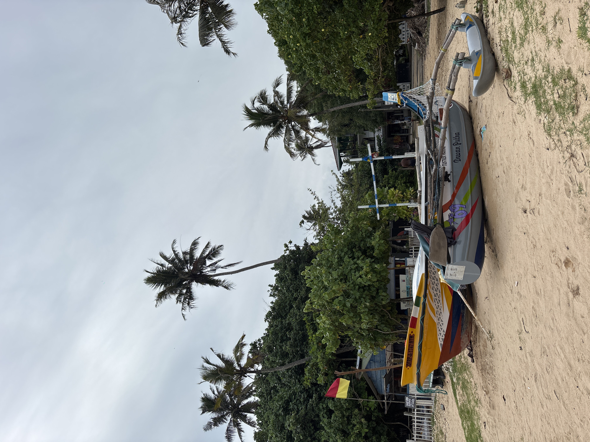 The image shows a beach in Weliweriya (SP), Sri Lanka. Several outrigger canoes rest on the sand near vegetation and palm trees. A yellow canoe with ornate designs and script leans against a grey canoe labeled "Dewan Putha" in script. Further away, another smaller blue and white canoe is also on the sand. Behind the boats are some buildings and trees. The sky is overcast.
